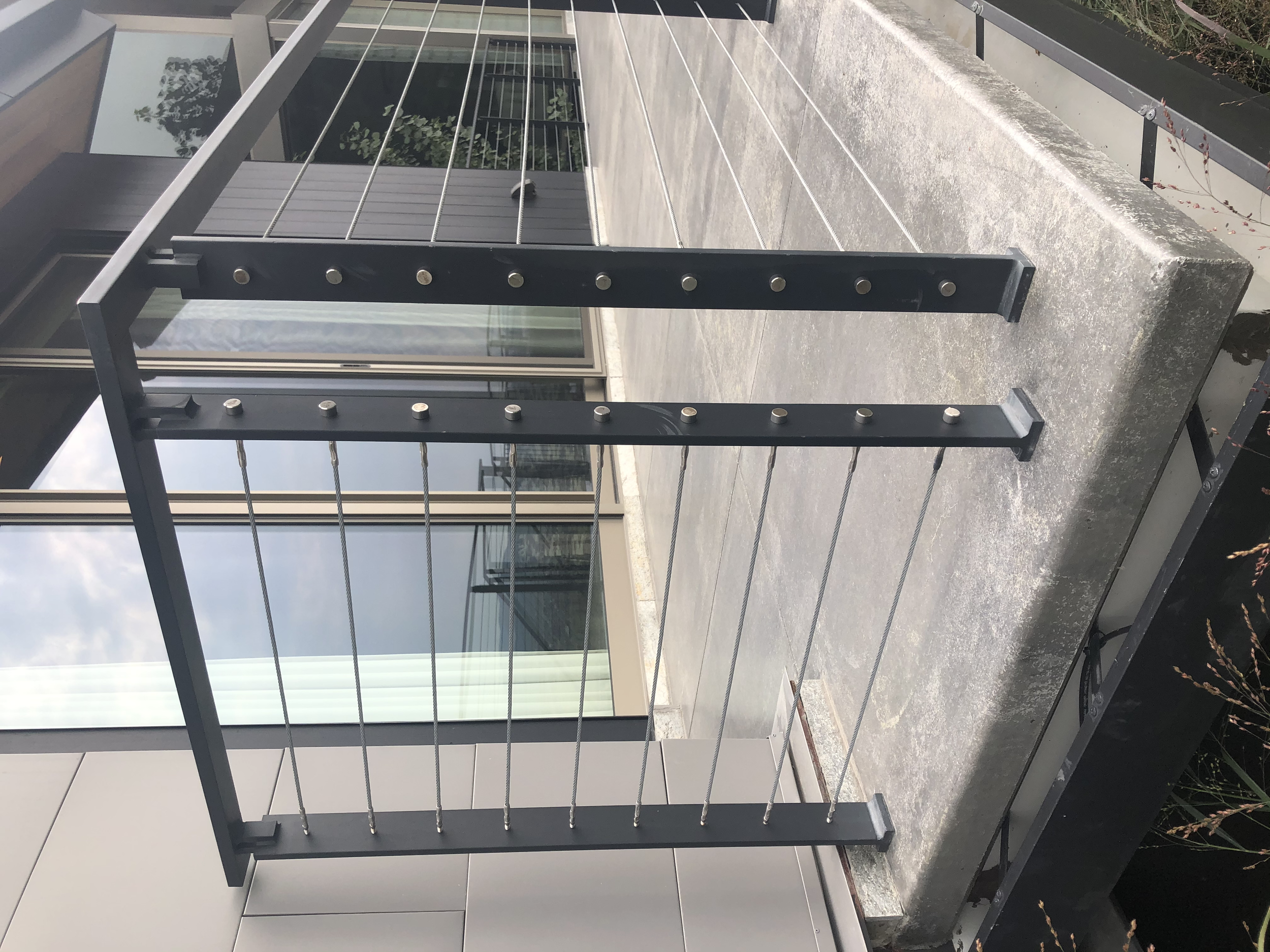 Close-up of a modern black metal railing with horizontal cables, installed on a concrete balcony outside a building with glass windows and doors.