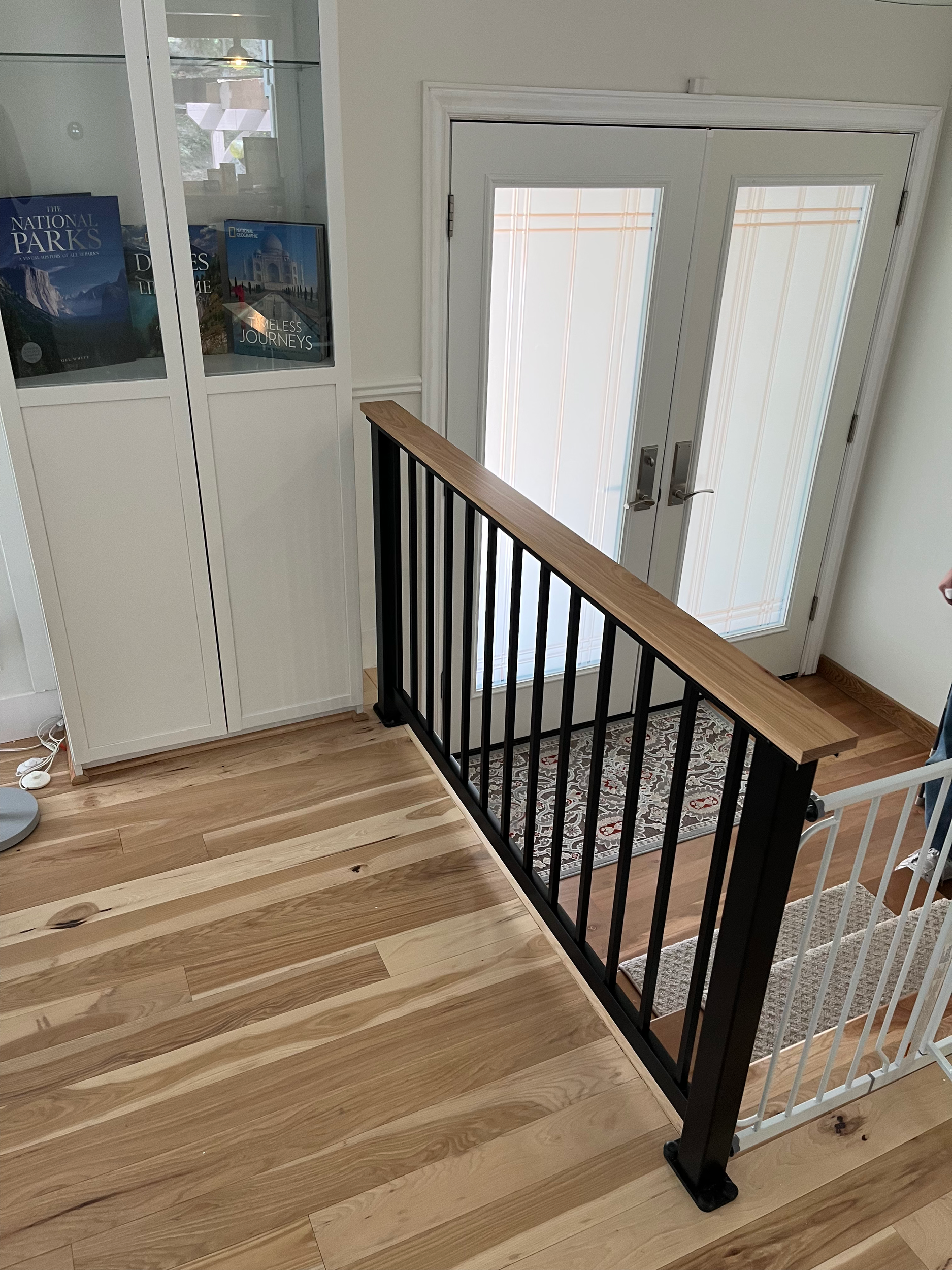 View of a stairway with a black metal railing, leading to a door with glass panels. To the left, a white cabinet with books inside, including titles about national parks and travels. The floor is made of light wood, and a patterned rug is partially v