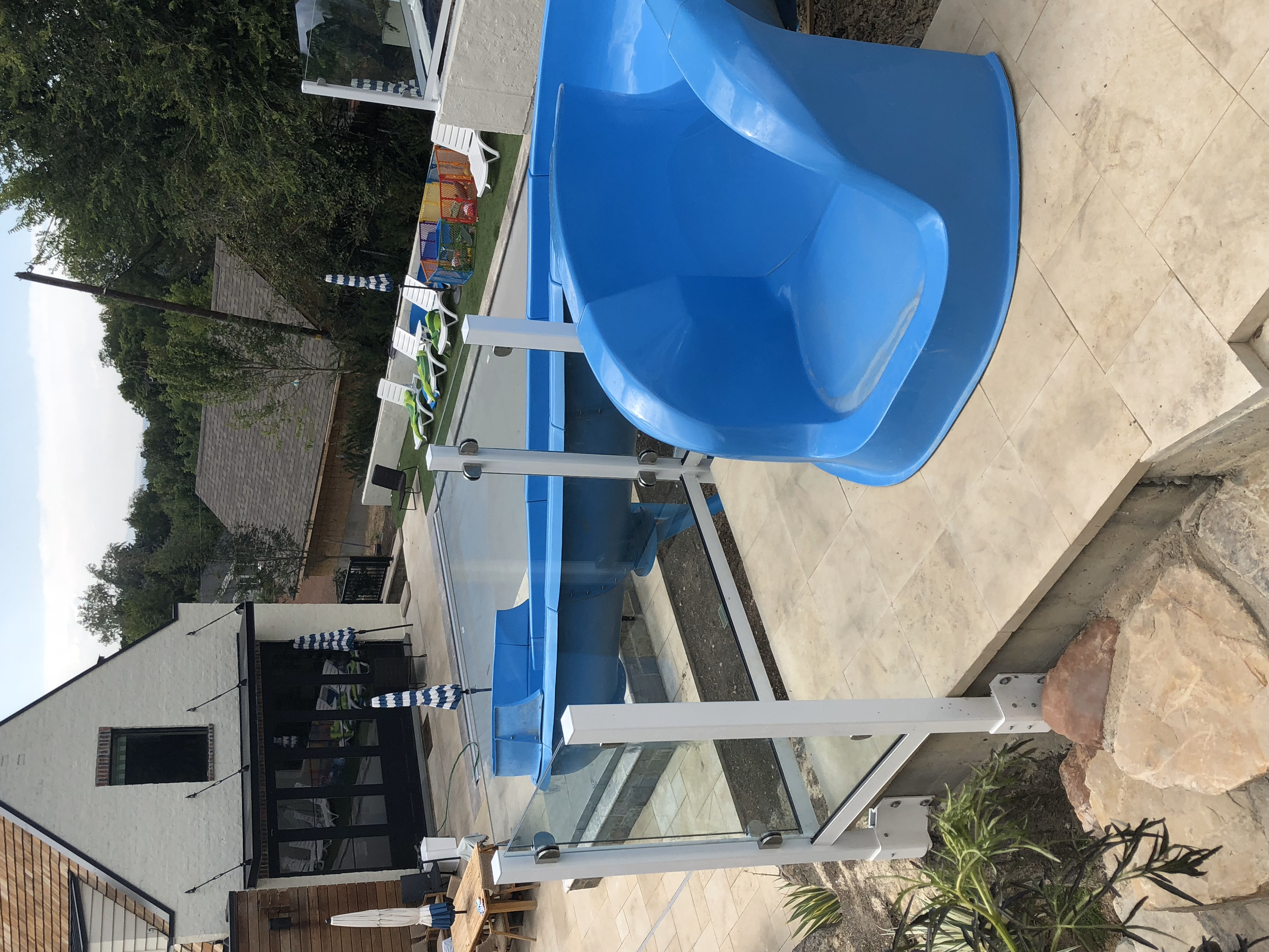 Backyard with a swimming pool and a blue waterslide, surrounded by lounge chairs, umbrellas, and a house with a white brick exterior and large glass railing systems. 