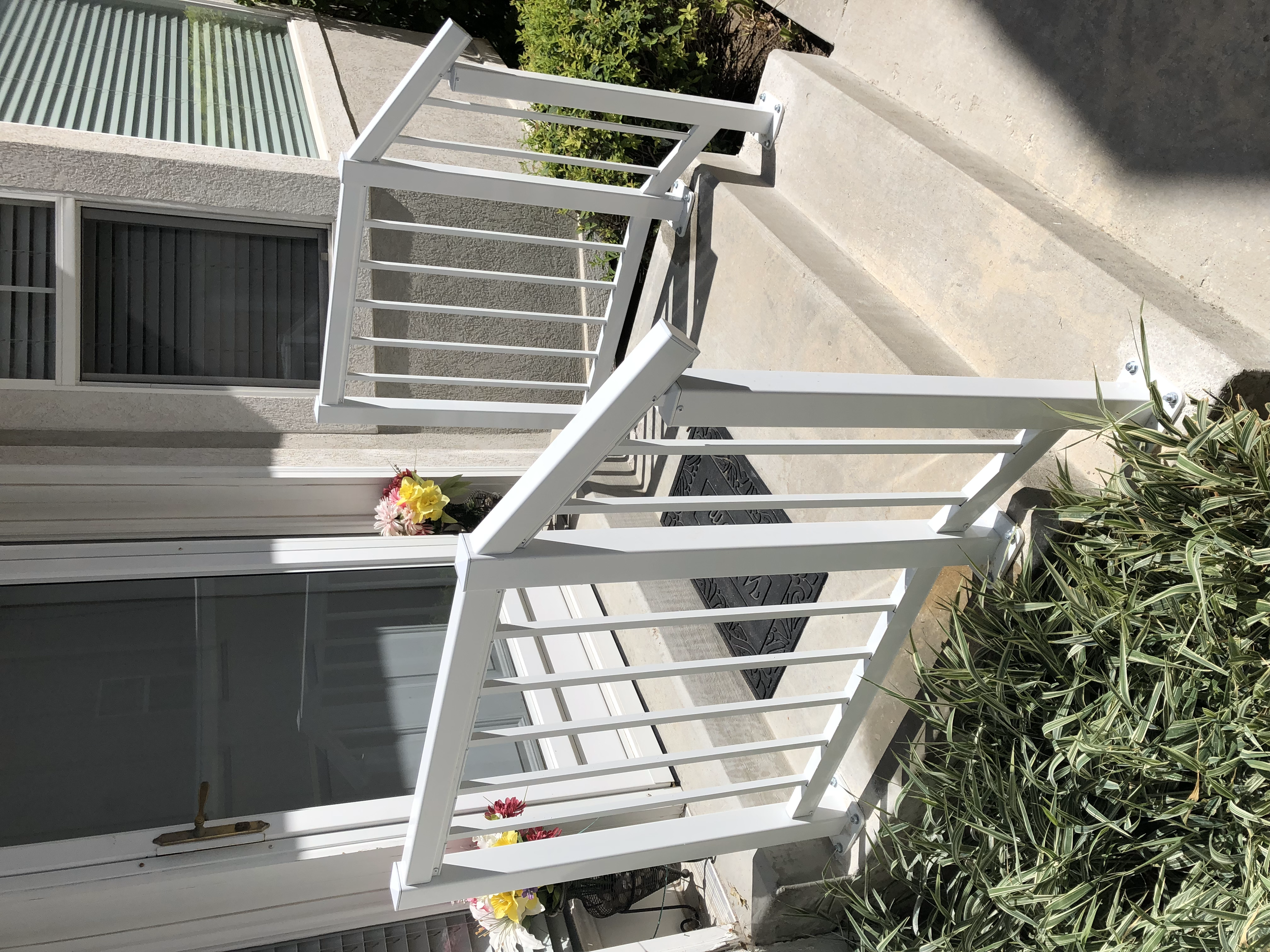 Front porch area with stairs, white railing, a sliding glass door, two windows with blinds, flower pots with pink and yellow flowers, and surrounding green bushes.