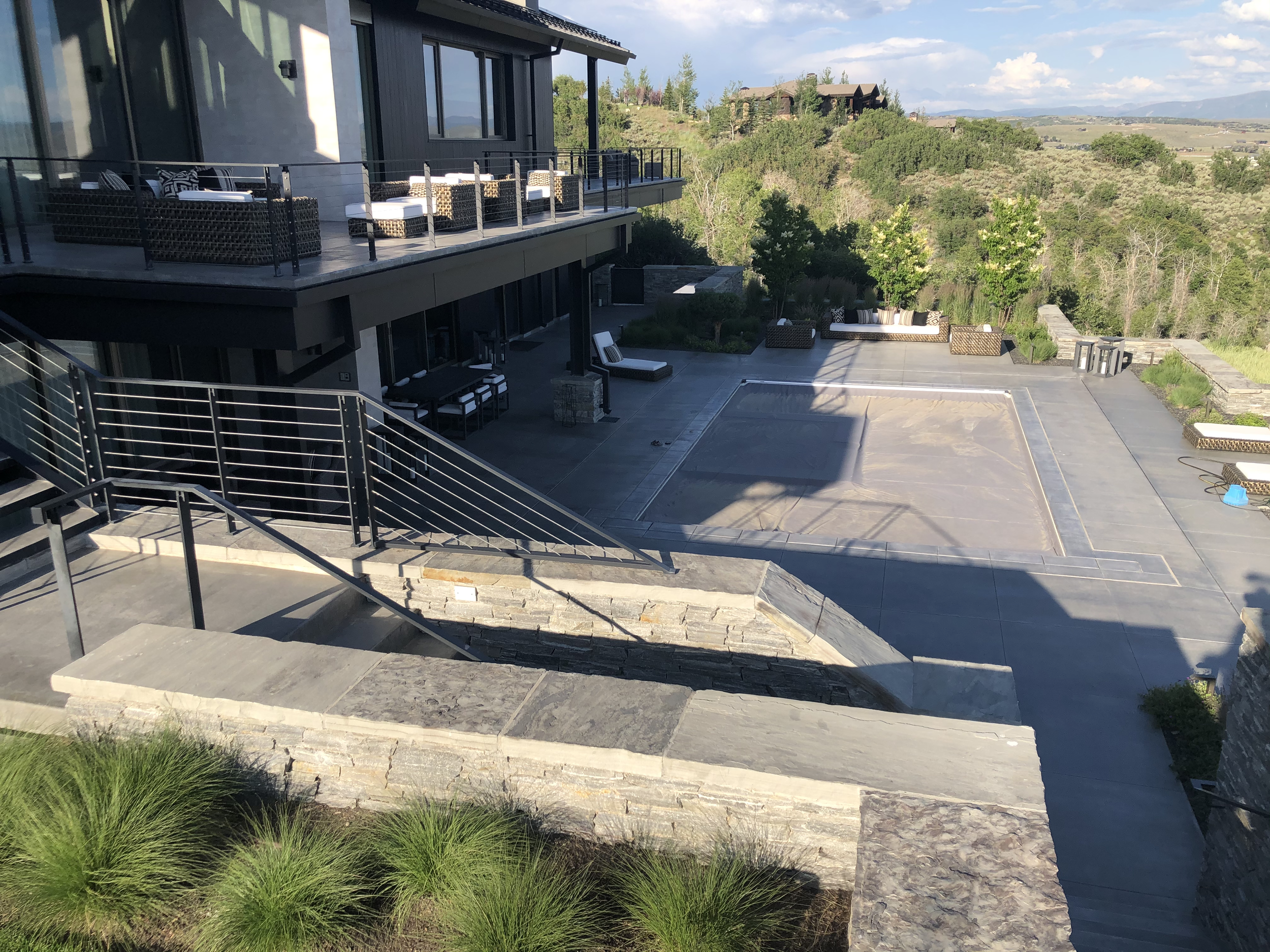 A spacious outdoor patio with modern outdoor furniture, including cushioned chairs and sofas, overlooking a green hilly landscape with scattered houses and trees.