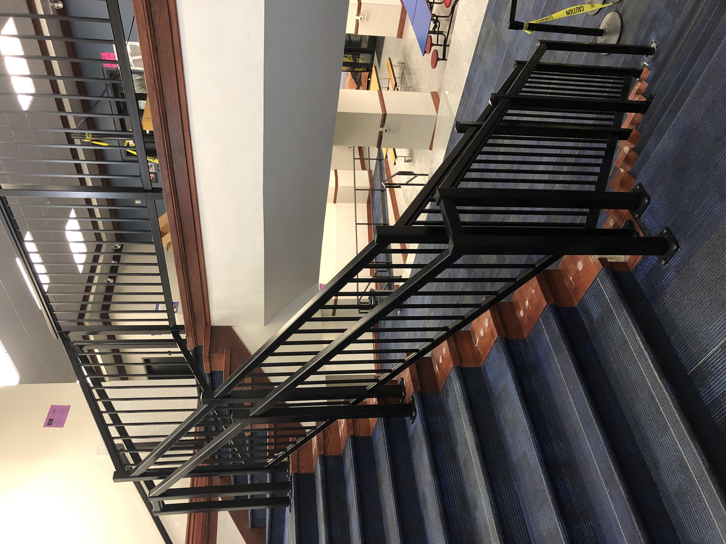 Inside view of a stairway with black metal railings, dark carpeted stairs, and a view of multiple floors with similar railings and a hallway with tables and chairs in the background.