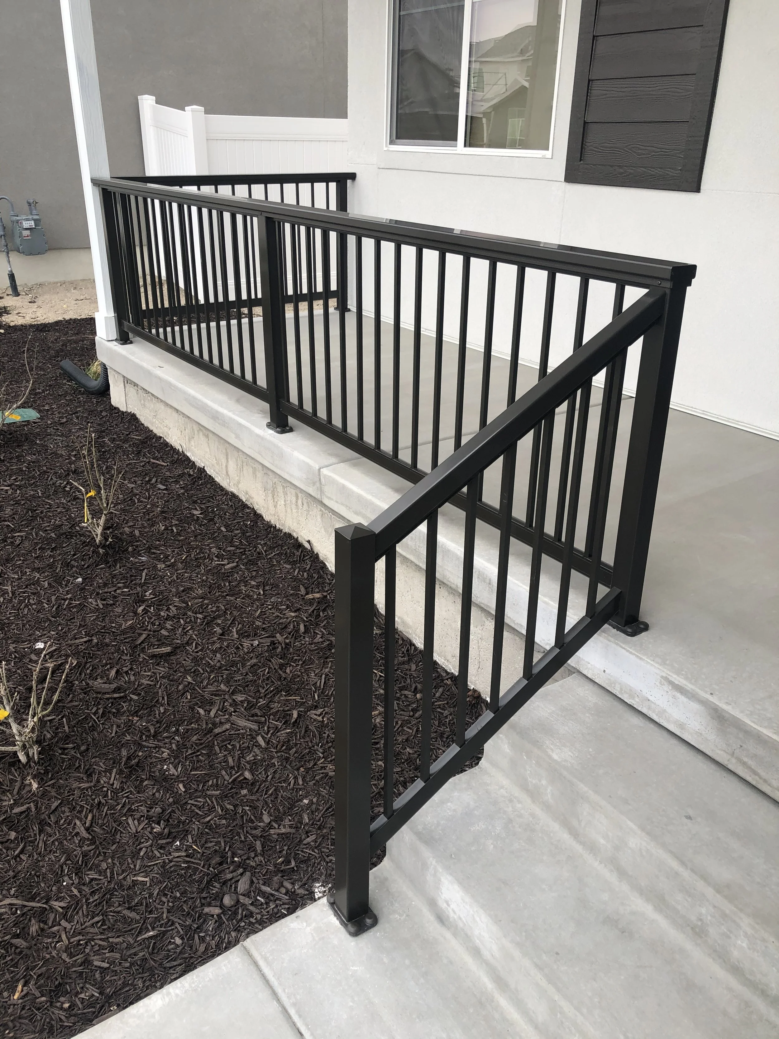 Black metal railing surrounding a small concrete porch area in front of a house with white walls, a window, and dark brown shutters.
