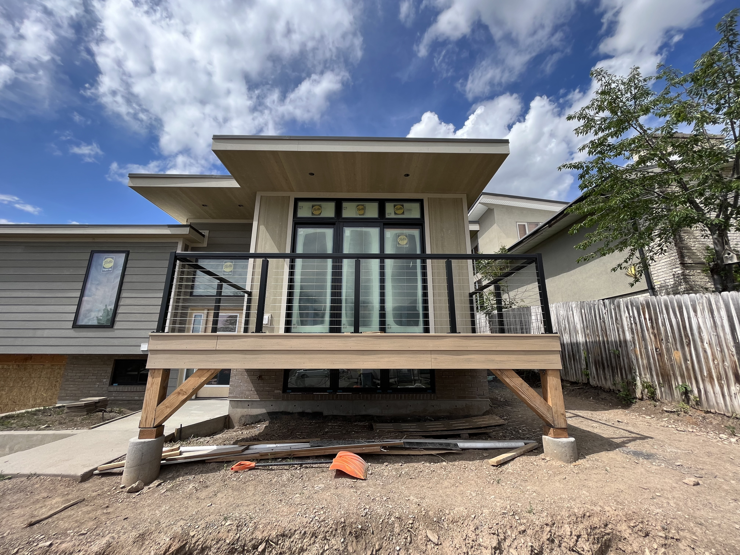 View of a modern house under construction with a wooden deck supported by wooden beams, large glass doors, siding, and a partially built balcony, set against a partly cloudy sky.