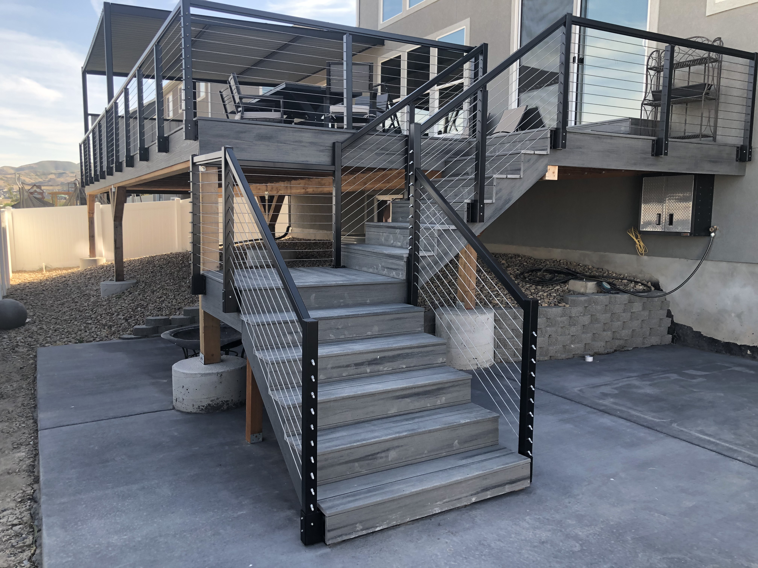 An outdoor balcony with a metal railing, wooden stairs, and a table with chairs. There is a house behind with windows and a door, and a mountain scenery in the background.