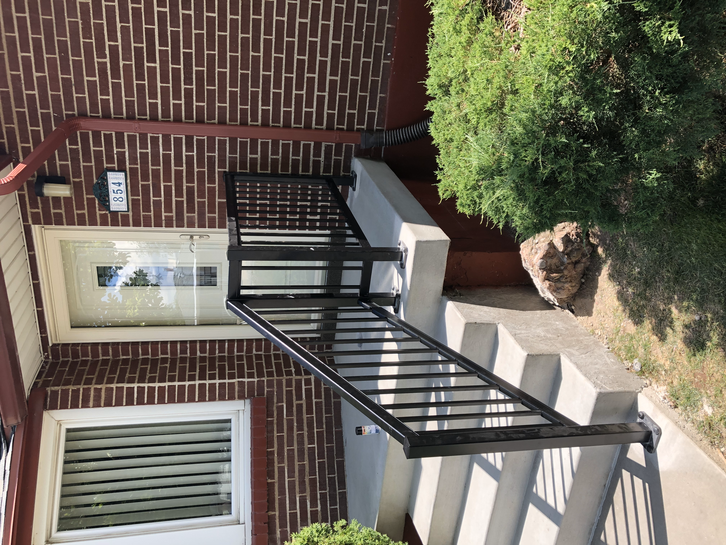 Exterior view of a front porch with concrete stairs, black metal railing, and a white door with a small glass window. There is a brick wall and window to the left of the door, a house number plaque with blue and white colors, a red downspout, and som