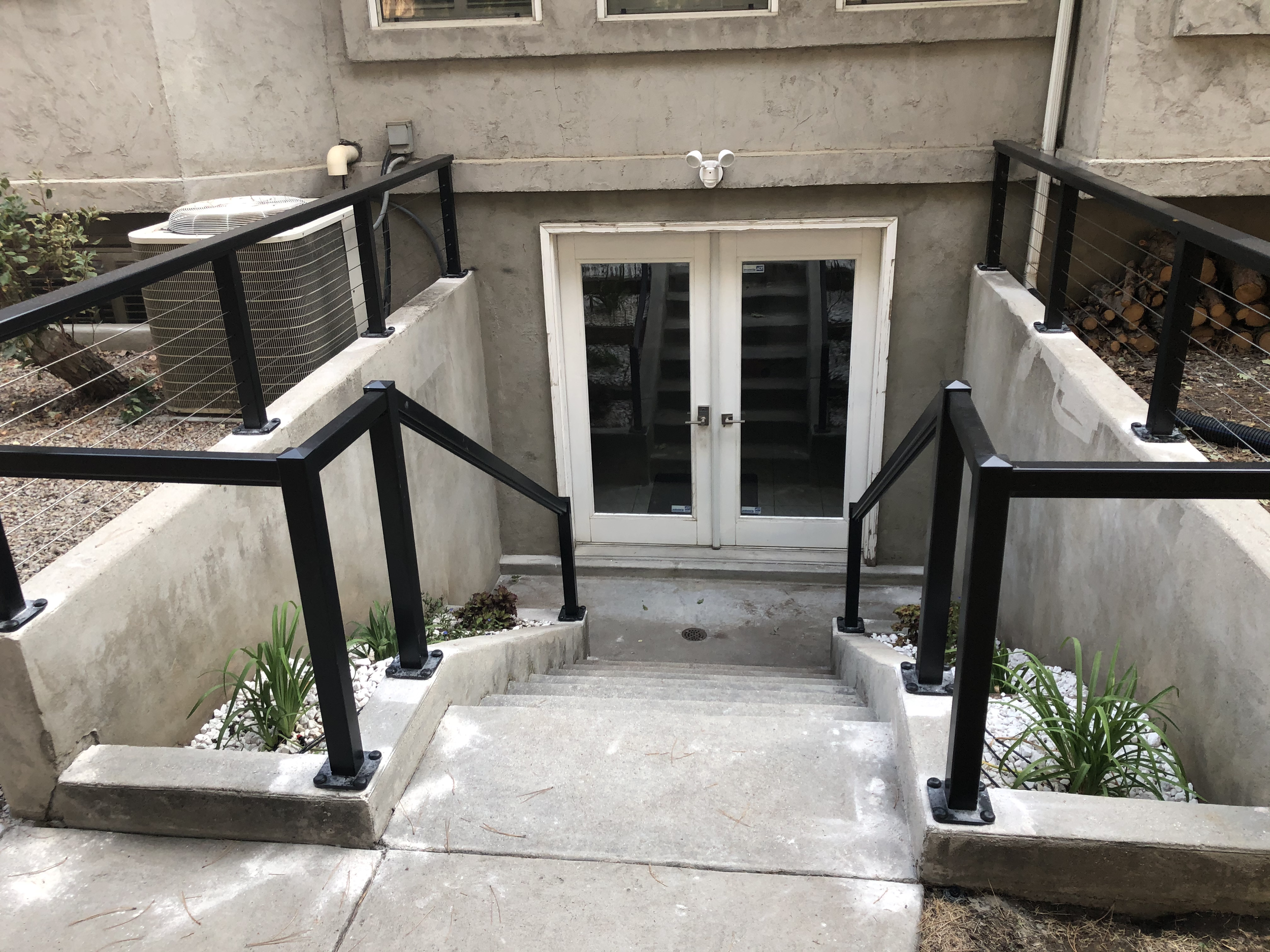 Concrete staircase leading down to a basement door with glass panels, black metal railings on both sides, an air conditioning unit on the left, a wood pile on the right, and surveillance cameras above the door.