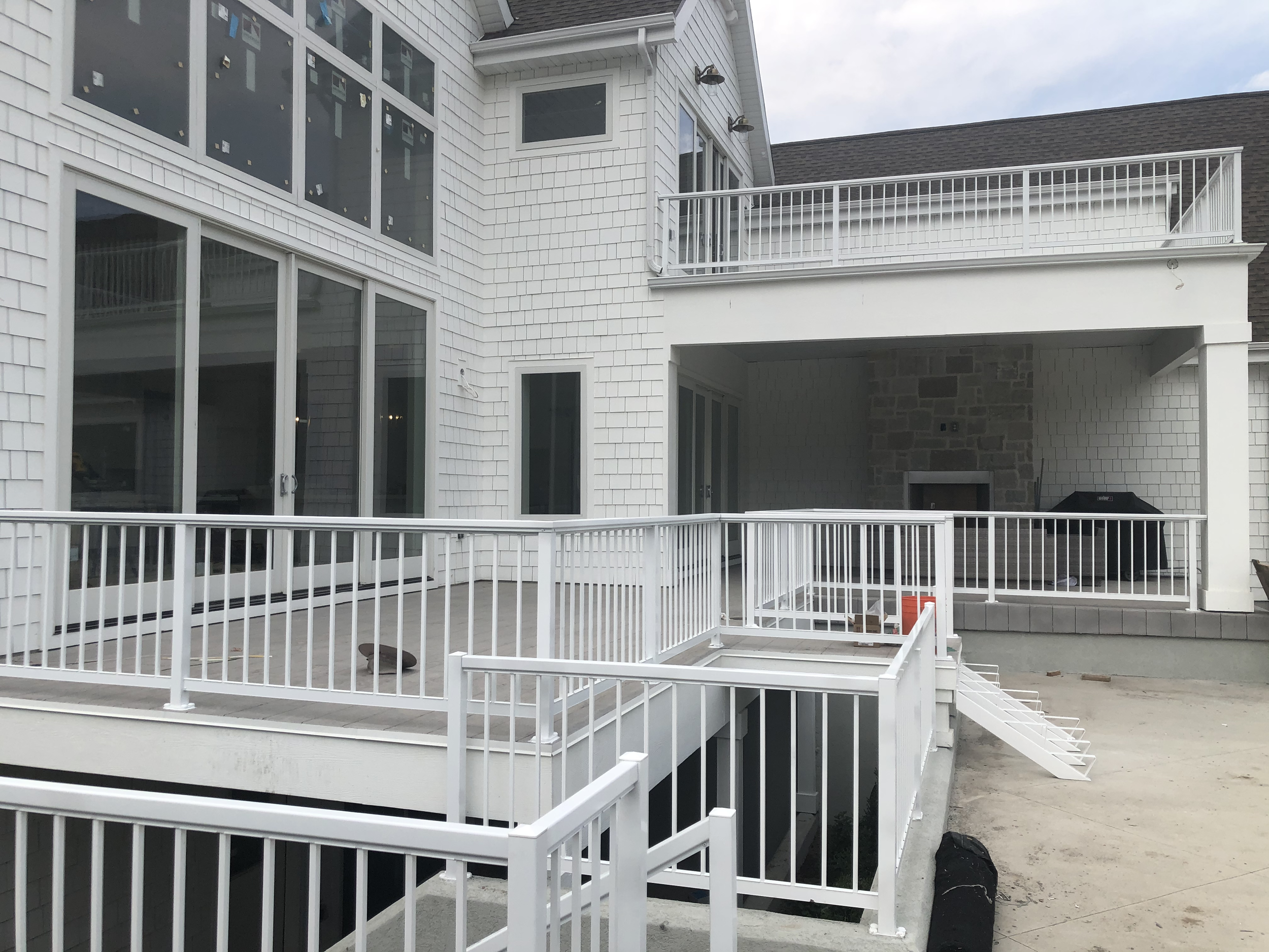 A multi-story house with white siding and large windows, featuring a spacious deck with white railings and an outdoor staircase, under construction with some building materials and tools visible.