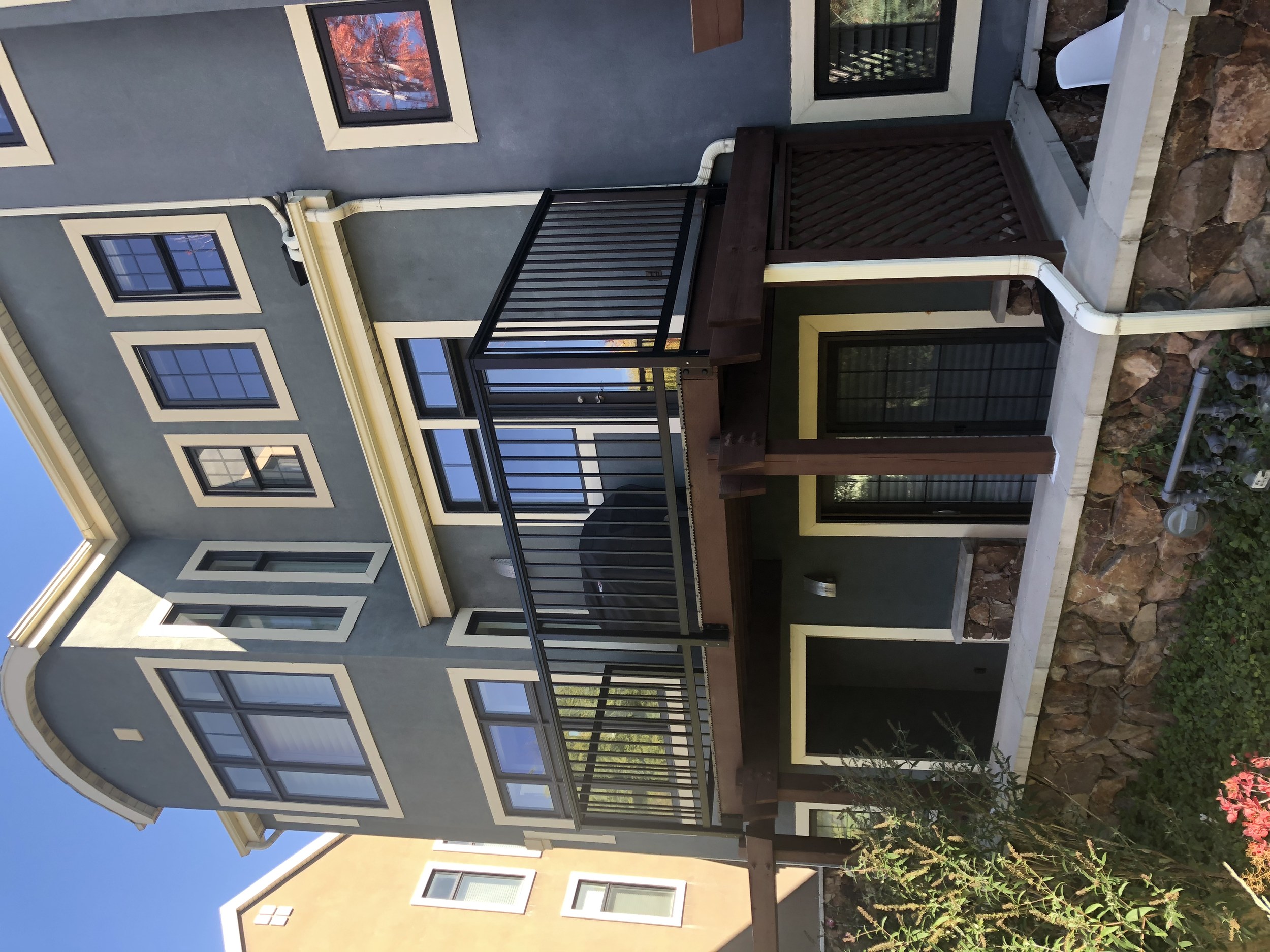 The image shows the exterior of a modern, multi-story house with a gray painted stucco finish and multiple windows. There is a balcony with a black metal railing on the second floor and a small area below with a stone foundation. The house has a wood