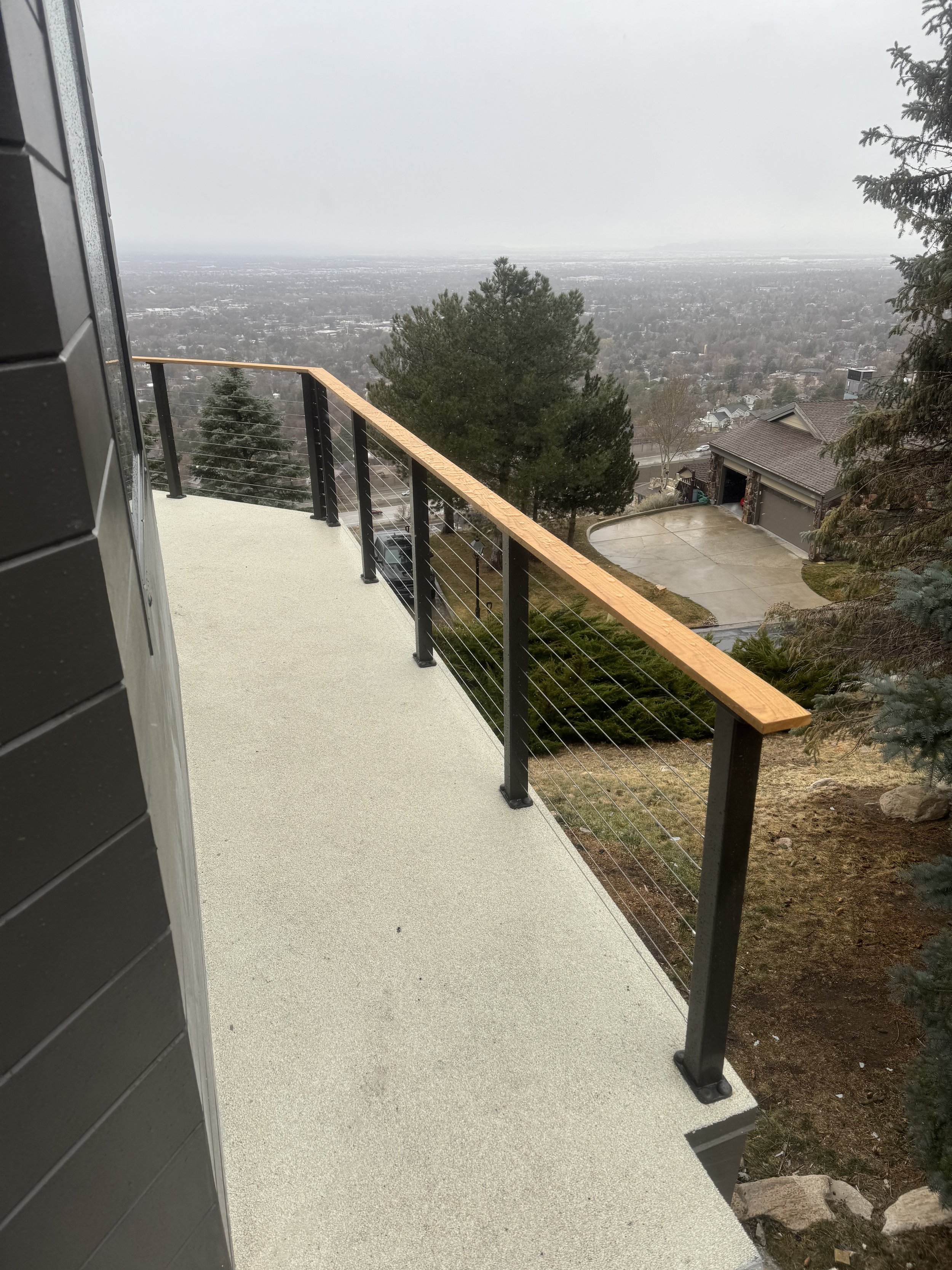 View from a balcony overlooking a cityscape with trees and houses. A black railing with a wooden top runs along the edge of the balcony, with a textured beige surface. The scene is overcast and slightly foggy.