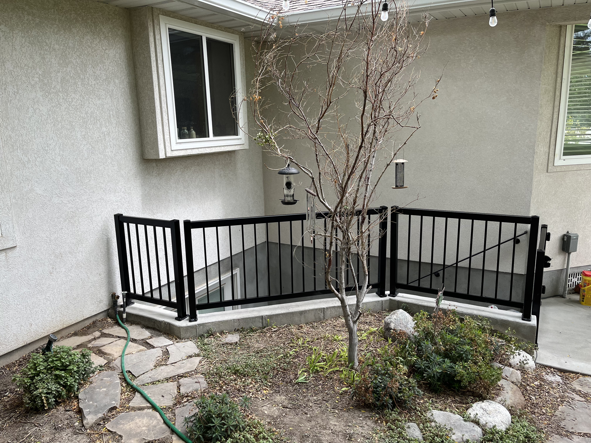 A backyard patio area with a small leafless tree, a black metal safety fence, and a garden with plants, rocks, and a garden hose on the ground.