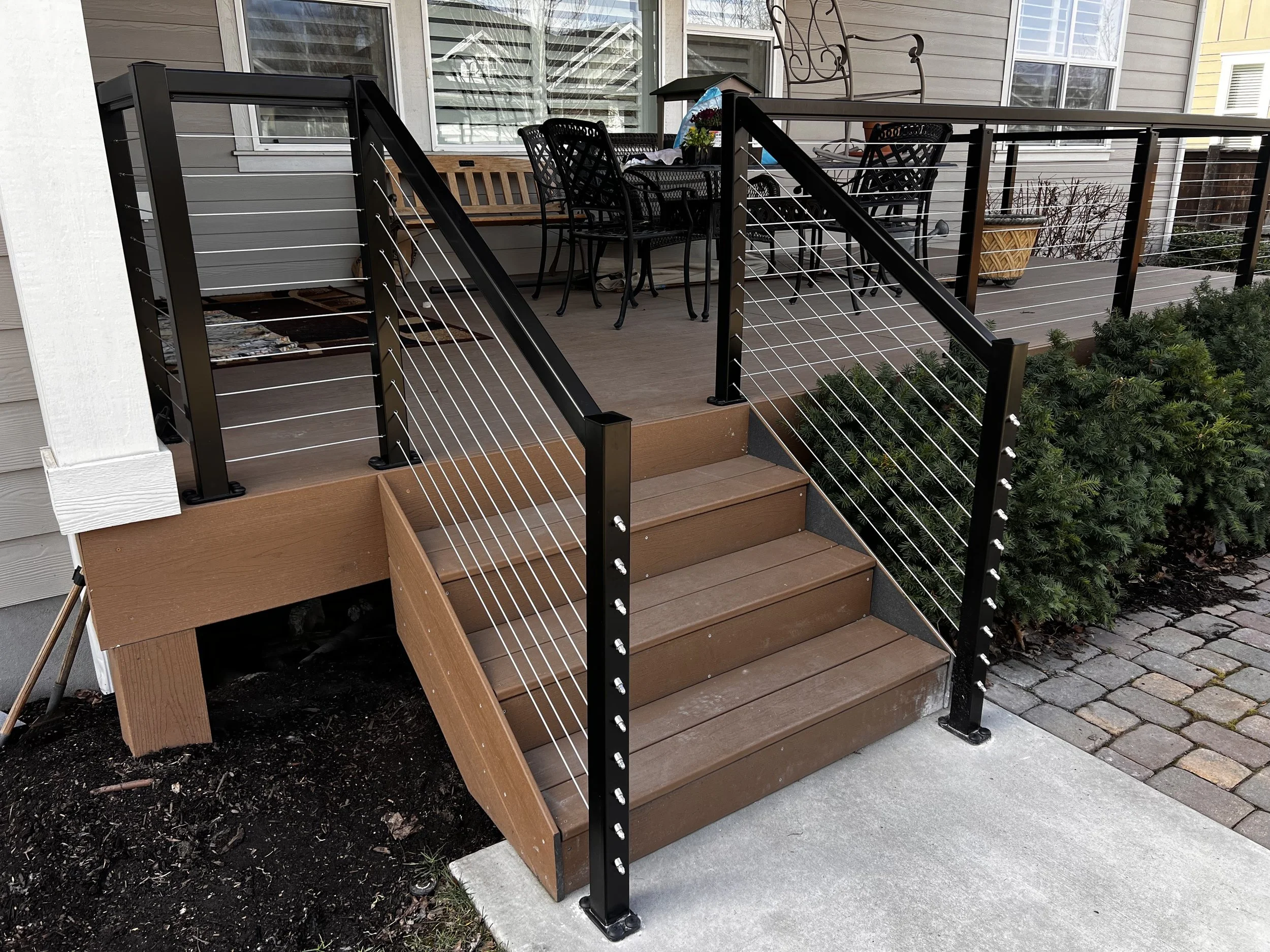 New wooden outdoor deck with metal and cable railings, accessible by wooden stairs, located beside a house with large windows, surrounded by bushes and concrete and brick pavement.