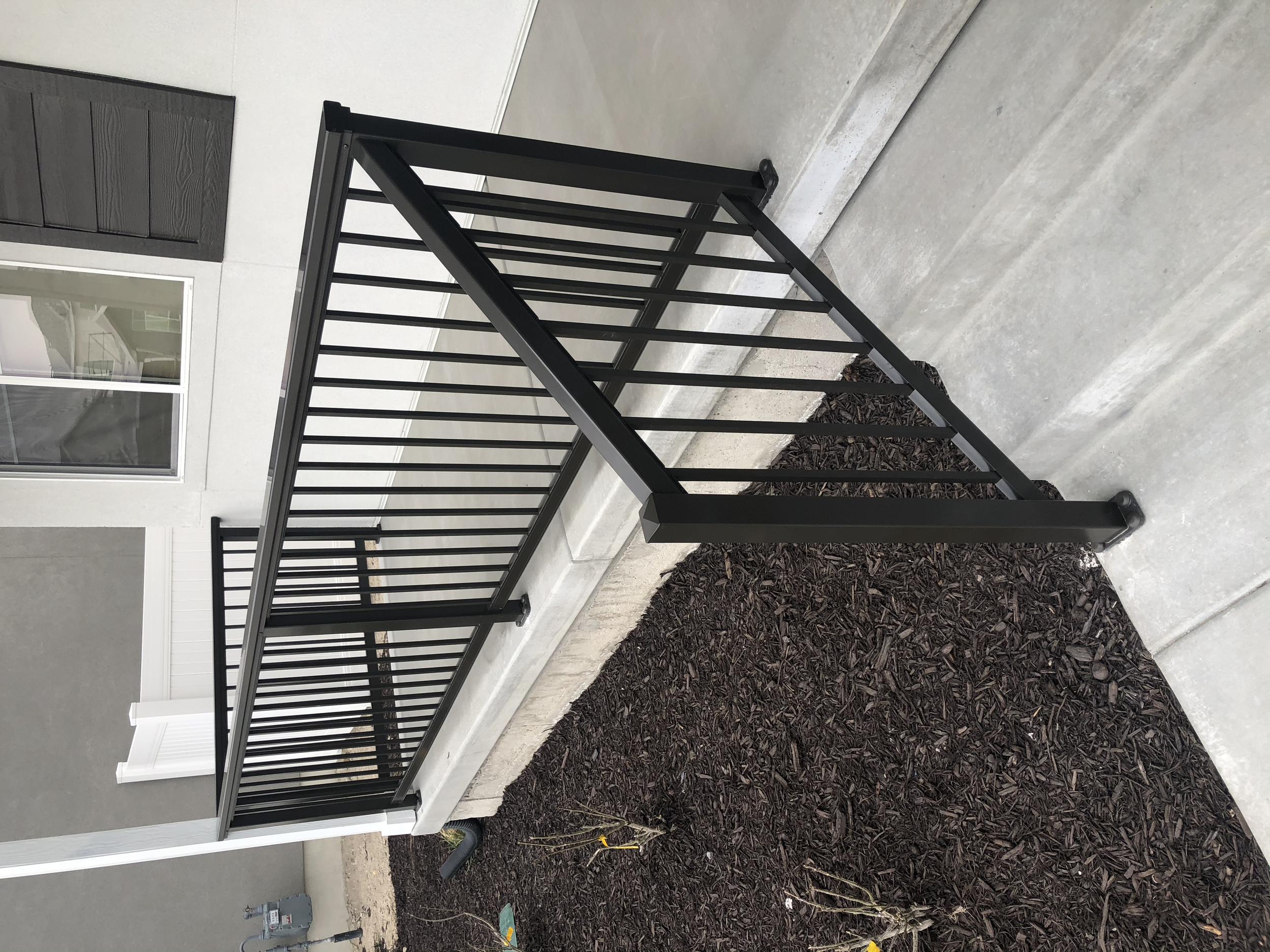 A black metal railing on a concrete porch outside a house with white and black exterior walls and a window with a black shutter.