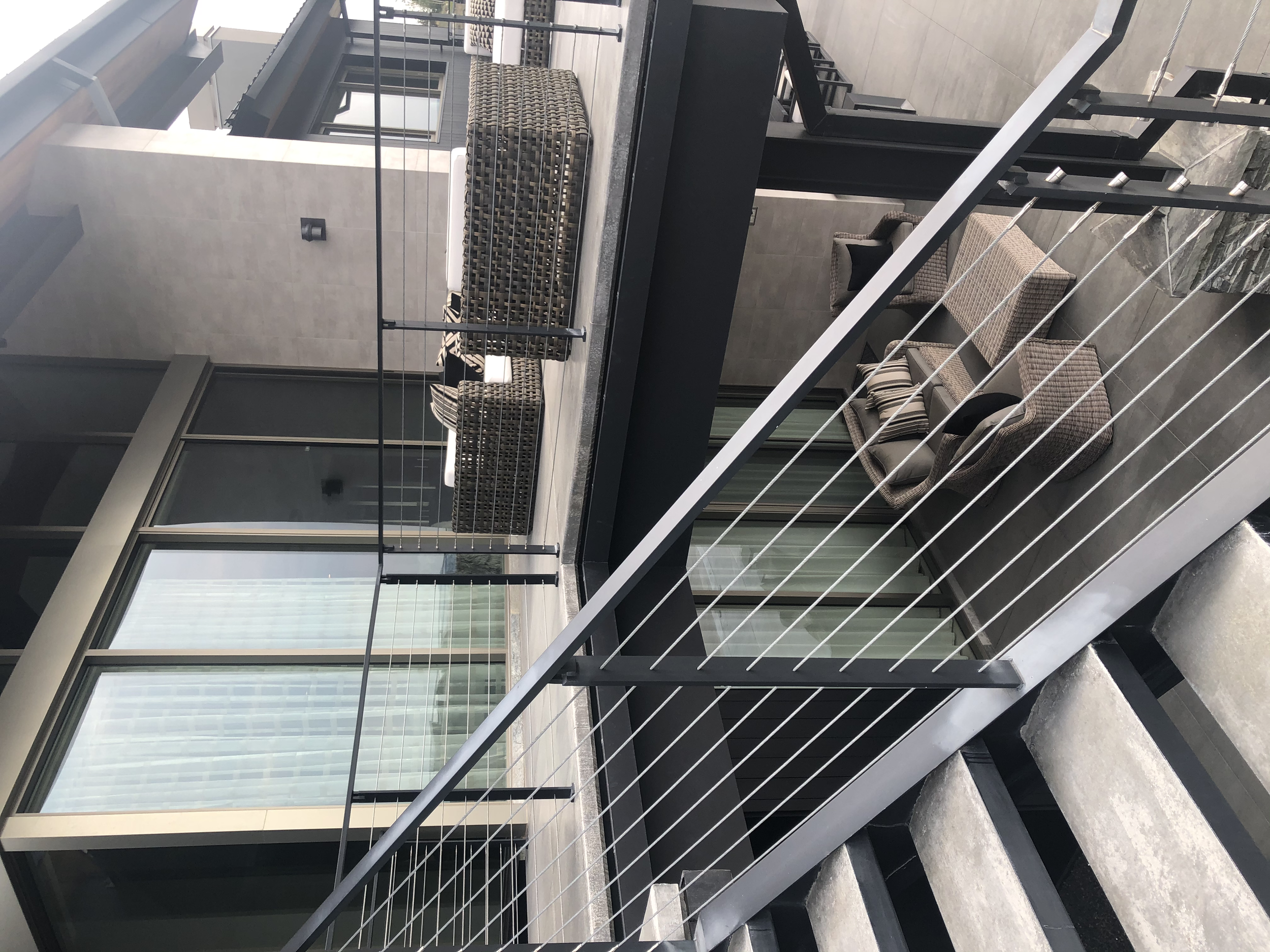 Exterior view of a modern building with a balcony that has wicker and cushioned outdoor furniture, large glass windows with curtains, concrete stairs, and a metal railing in an urban setting.