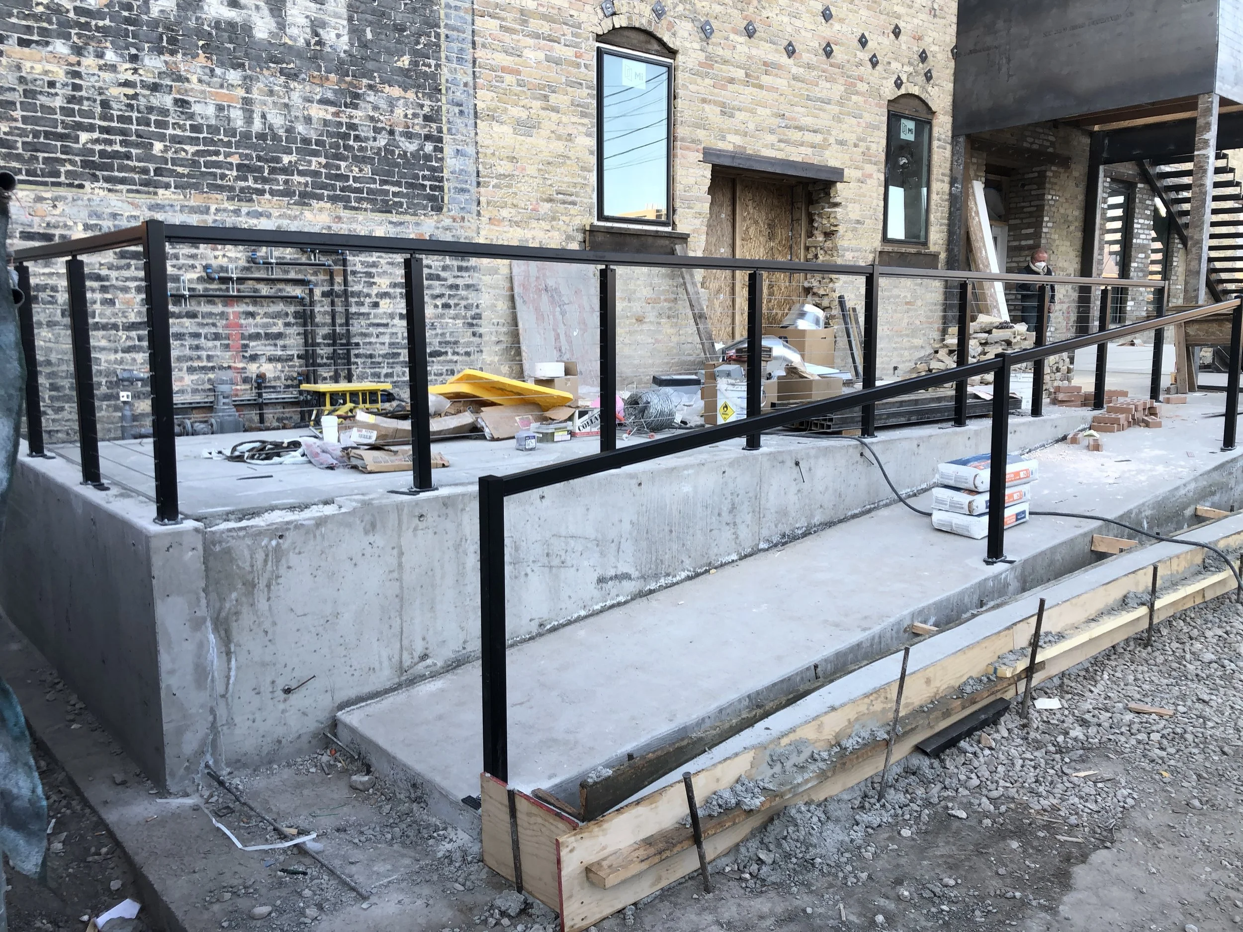Construction site with a concrete foundation, brick walls, and black metal railing. Construction tools, materials, and debris are scattered around, with wooden forms set up for future concrete pouring.