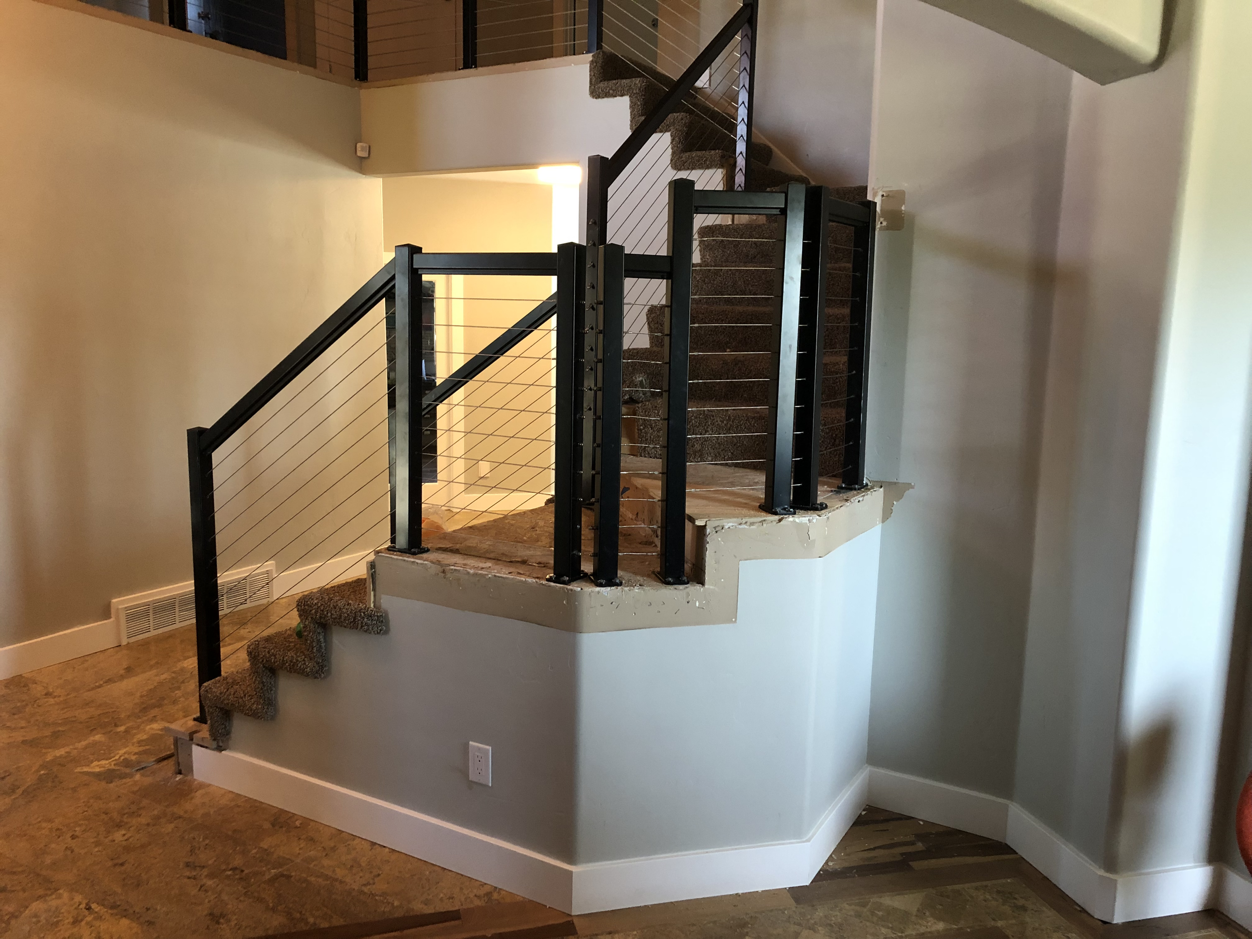 Interior view of a staircase under construction with black metal railings, wooden steps covered with carpet, and a partial view of a balcony with a metal railing. The staircase wraps around a corner and is situated next to beige and white walls, with