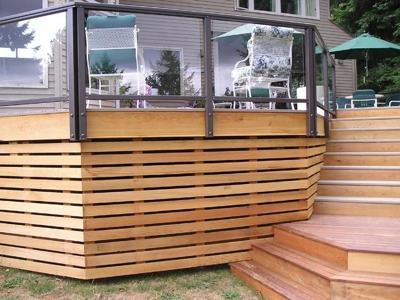 Wooden deck with glass railing and outdoor furniture, including chairs and an umbrella, attached to a house with siding.