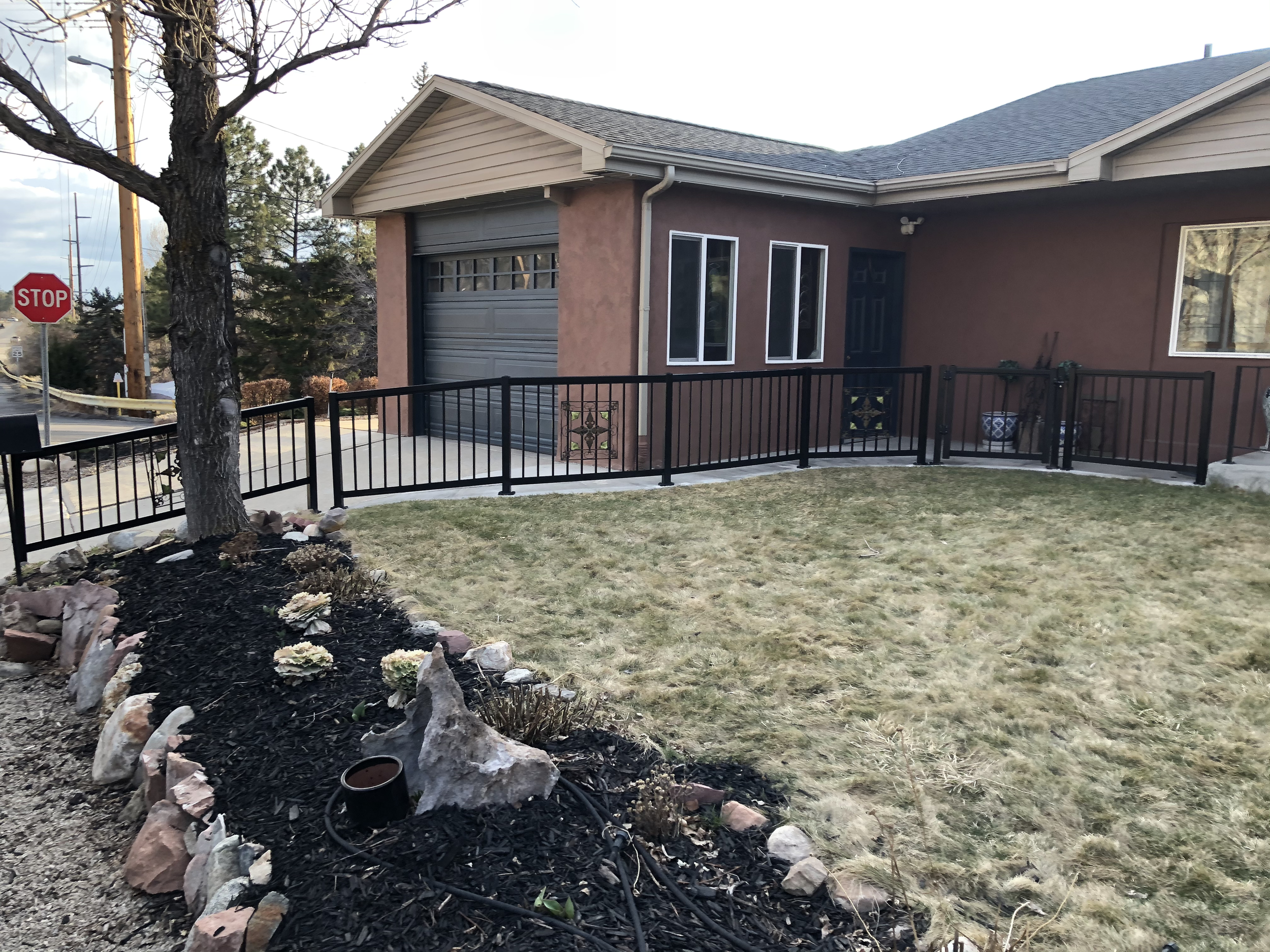 A residential house with a brown exterior and a gray garage door. There is a small fenced yard with a tree and a garden bed with rocks and plants in the foreground. A stop sign is visible on the street corner to the left.