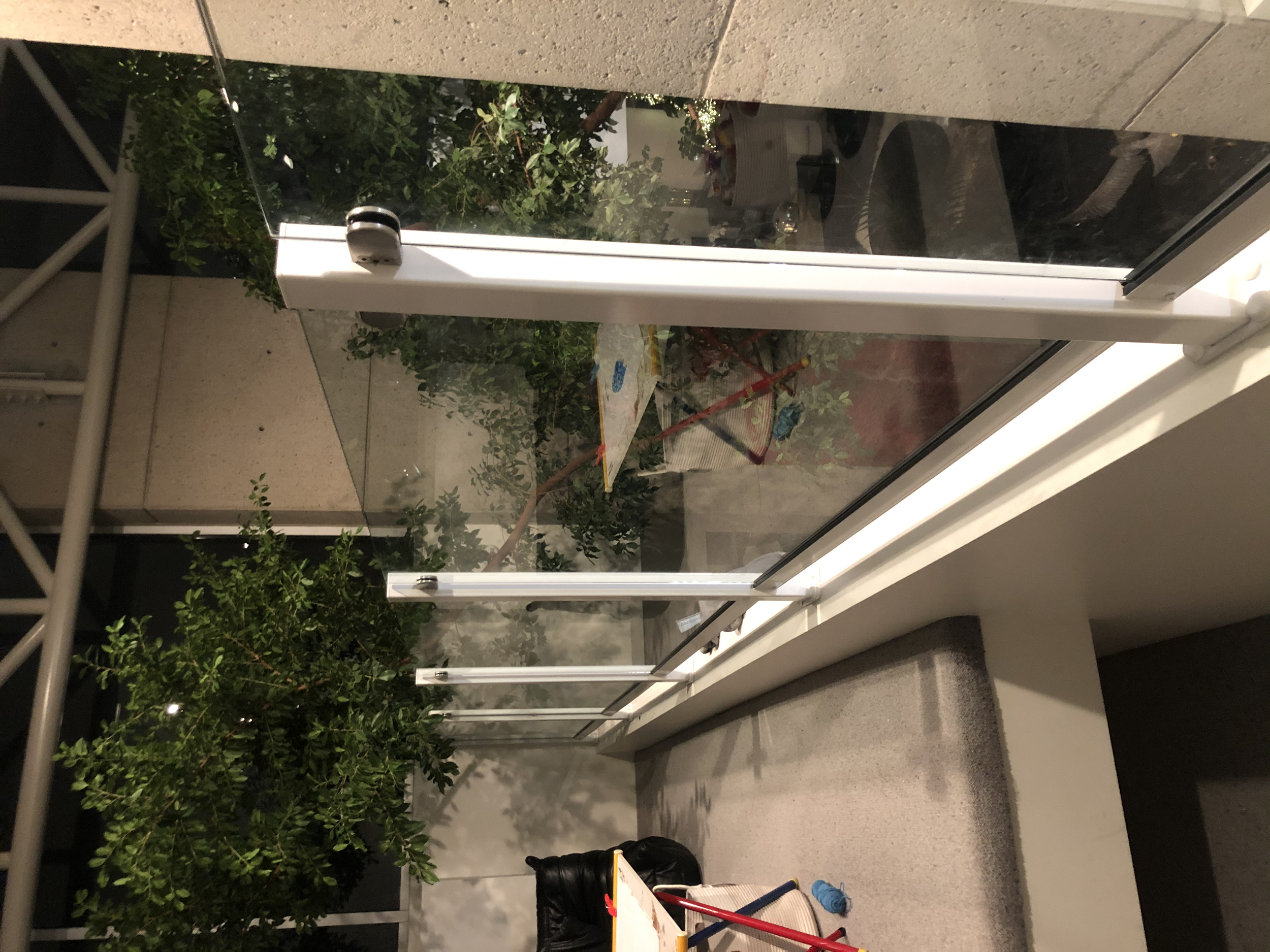 View of a glass safety barrier on a balcony or terrace, with foliage composed of green leaves behind it, nighttime setting.
