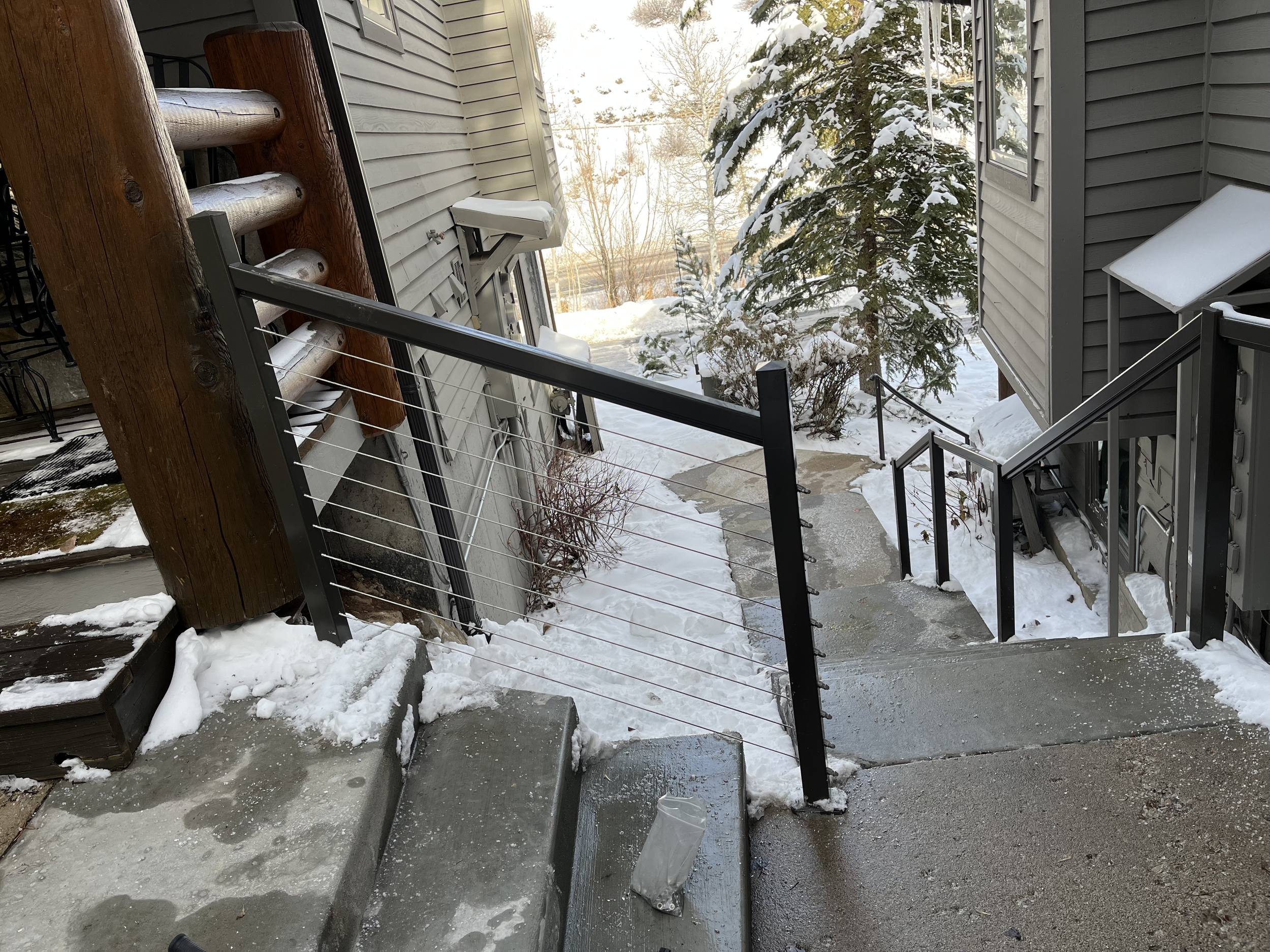 Snow-covered outdoor staircase with glass and wood railing, surrounded by snow and trees, houses in background.