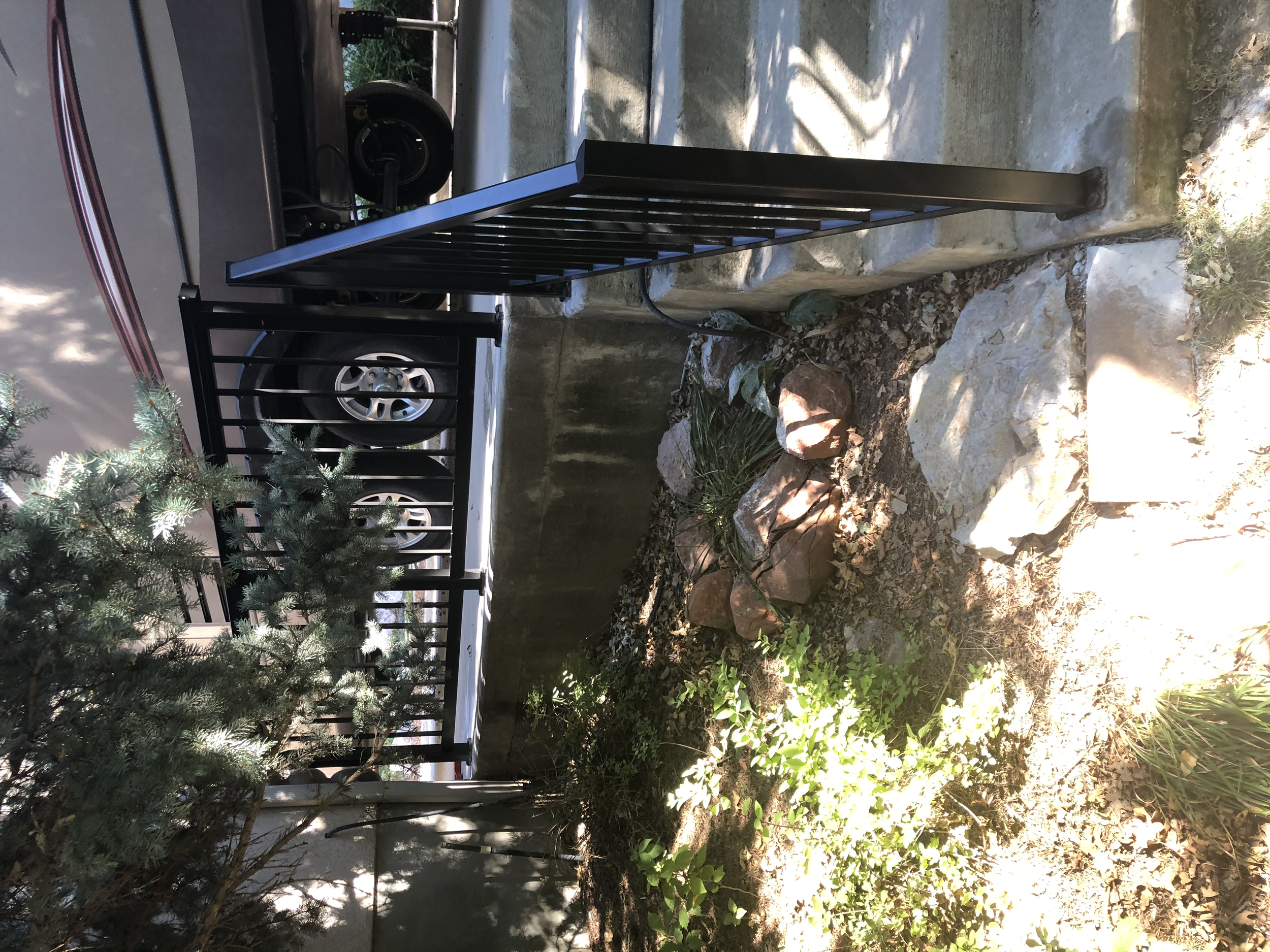 A set of concrete stairs with black metal handrails leading up to a porch, surrounded by greenery and rocks. A trailer with tires is visible at the top of the stairs. The porch is shaded with sunlight filtering through nearby trees.