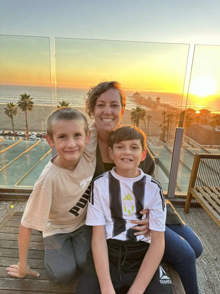 A woman and two boys smiling on a balcony overlooking a beach at sunset with palm trees and pier in the background.