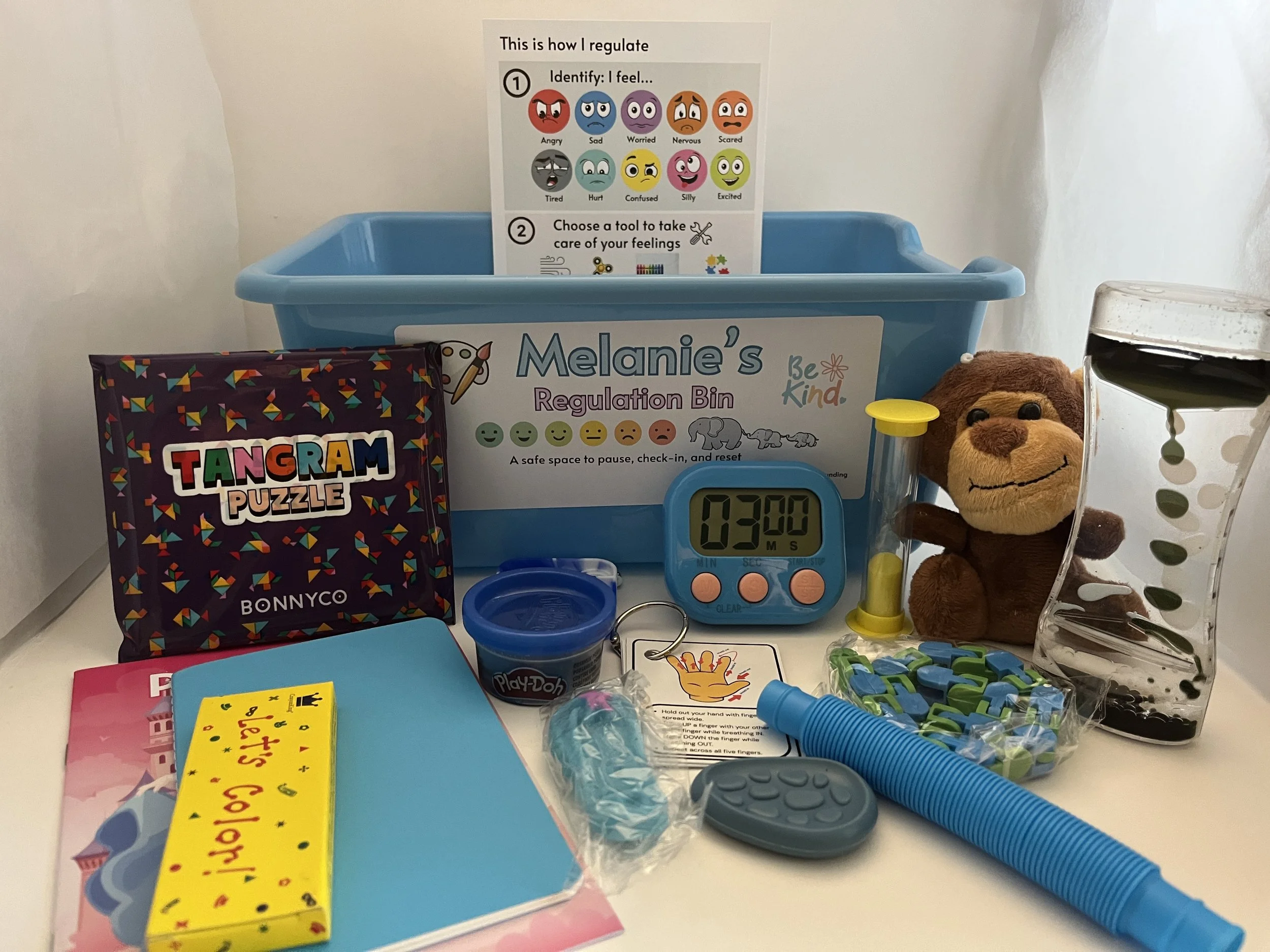 A collection of children's toys and educational materials on a white surface. Includes a blue regulation bin labeled 'Melanie's Regulation Bin,' a Tangram Puzzle box, a colorful scratch art book, a Play-Doh container, a digital timer showing 3 minutes, toy monkey, a small potted plant, and various small toys and game pieces.