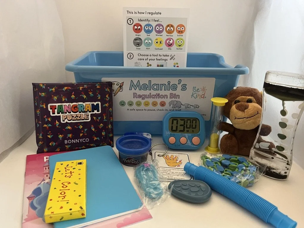 A collection of items on a white surface, including a blue regulation bin labeled Melanie's, a Tangram Puzzle, a pink planner, a yellow sticky note pad, a blue pencil, a blue timer displaying 03:00, a plush monkey toy, a small container of Play-Doh, a clear plastic container filled with colorful links, a gray device, and a black and clear water dispenser with a leafy design. There is also a printed emoji chart pinned inside the bin with the title 'This is how I regulate' and instructions to identify feelings and choose a tool for feelings.