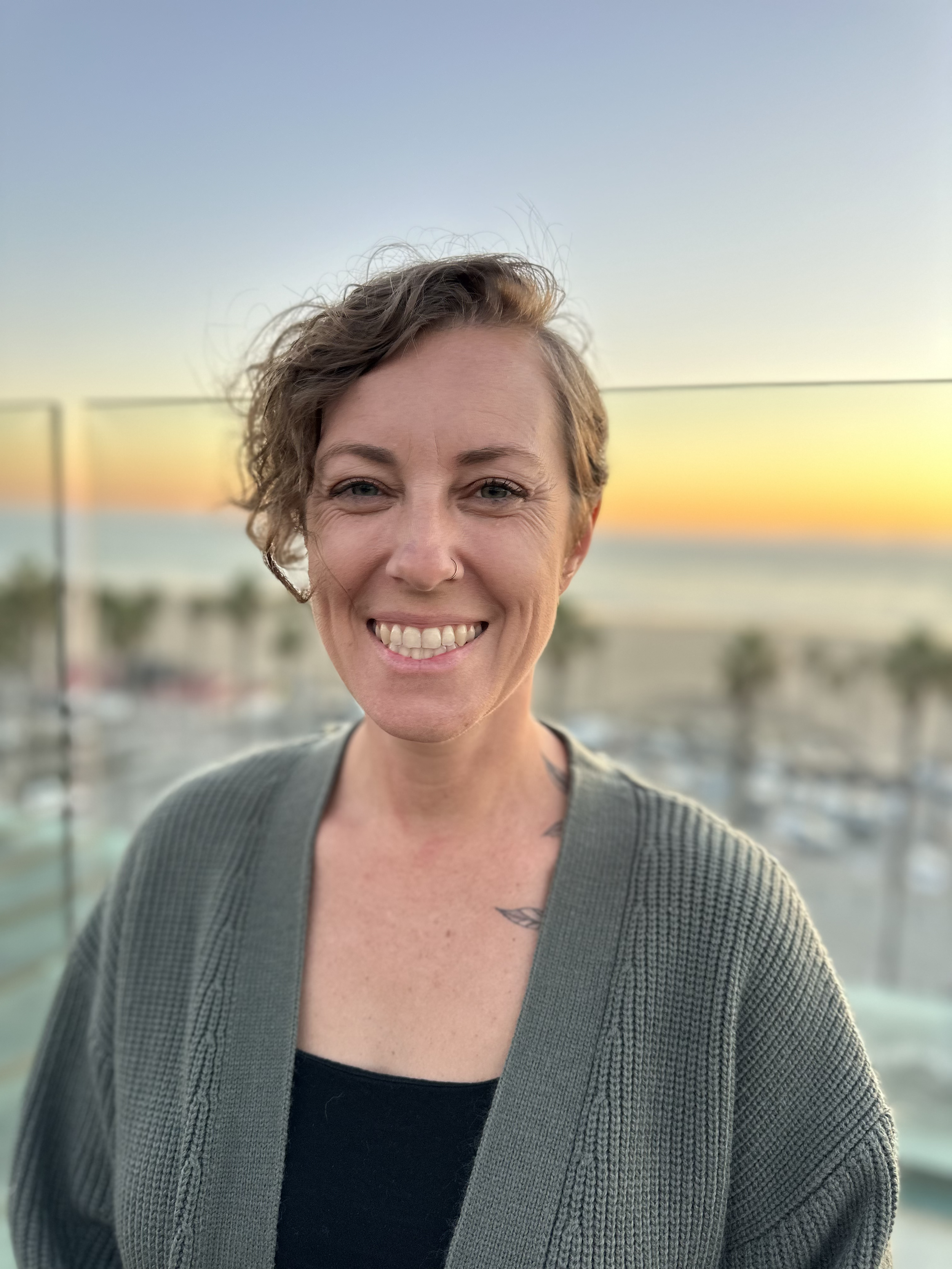 A woman with short curly hair smiling at sunset, standing near a body of water with a railing in the background.