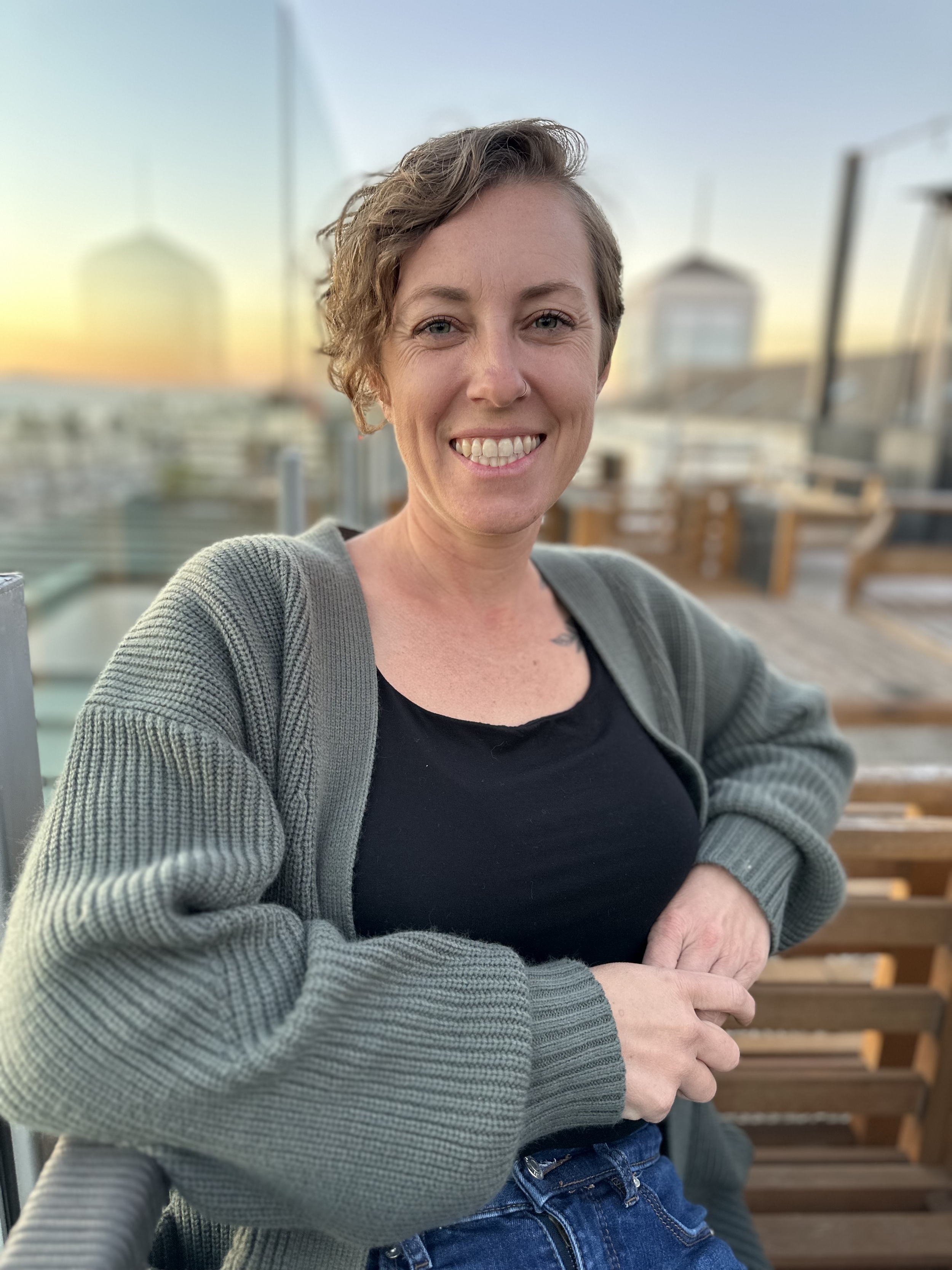 A woman with short curly hair and a nose ring smiling, wearing a black top and gray cardigan, sitting outdoors on a wooden bench during sunset.