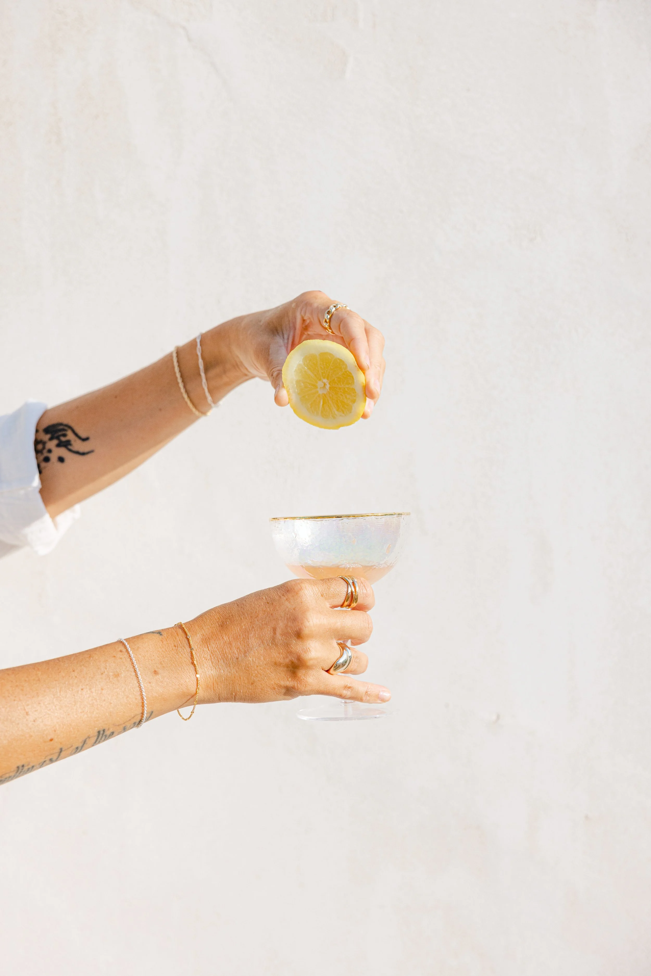 A person's hand holding a lemon half over a cocktail glass, with tattoos, jewelry, and a white wall background.