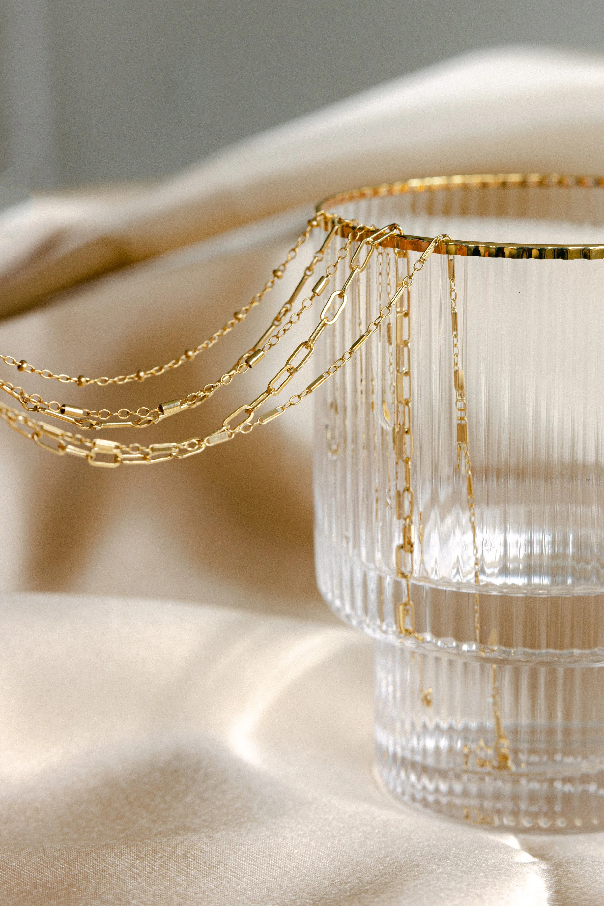 Close-up of gold jewelry necklaces draped over a glass container on a light-colored surface with a soft cloth in the background.