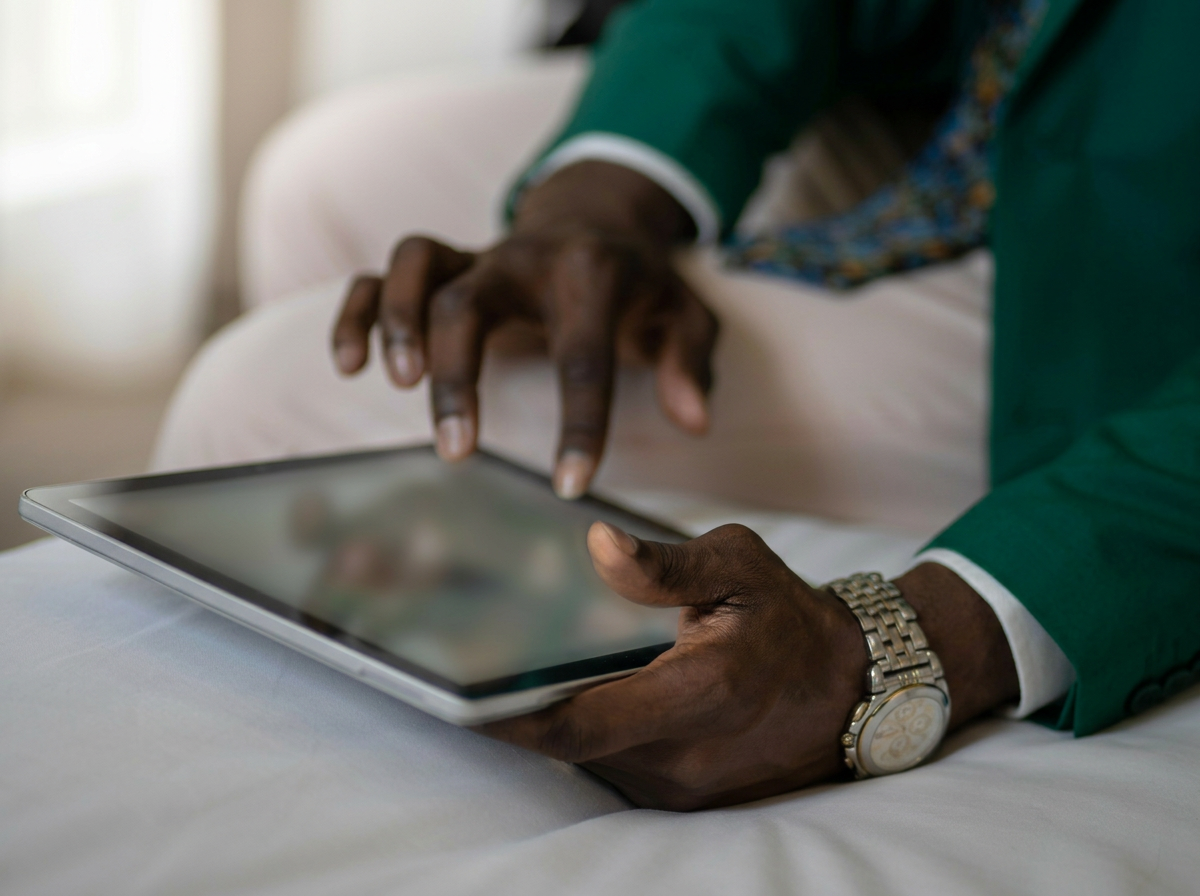 Close-up of a person with dark skin using a tablet, wearing a green blazer, white shirt, and a chunky silver watch, sitting on a bed.