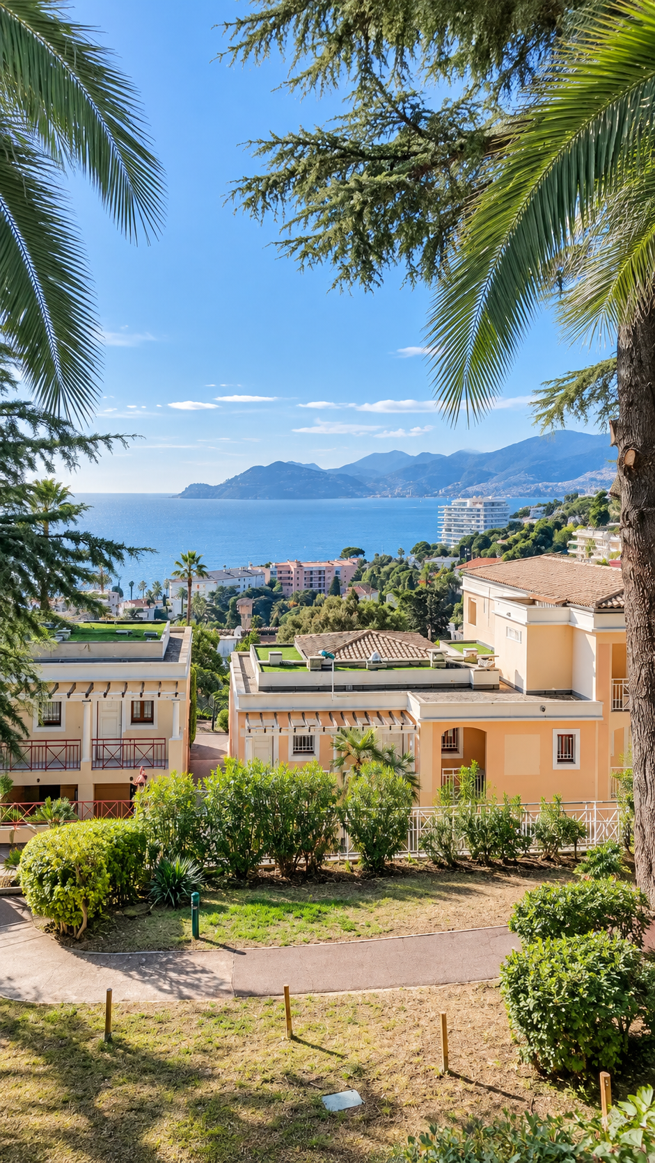 Vue panoramique d'immeubles résidentiels en bord de mer, avec la mer et des montagnes en arrière-plan, encadrée d'arbres et de végétation luxuriante, ciel bleu ensoleillé.