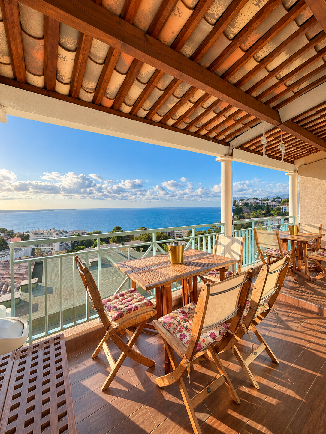 Balcon avec tables et chaises en bois, vue sur la mer et le ciel avec des nuages, bâtiment et végétation en arrière-plan, sous un plafond en bois avec des tuiles.