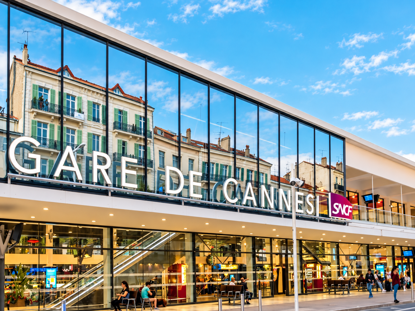 Vue extérieure de la gare de Cannes avec le nom écrit en lettres blanches, bâtiment moderne avec façade en verre, plusieurs personnes assises et marchant devant, en arrière-plan un ciel bleu avec quelques nuages.