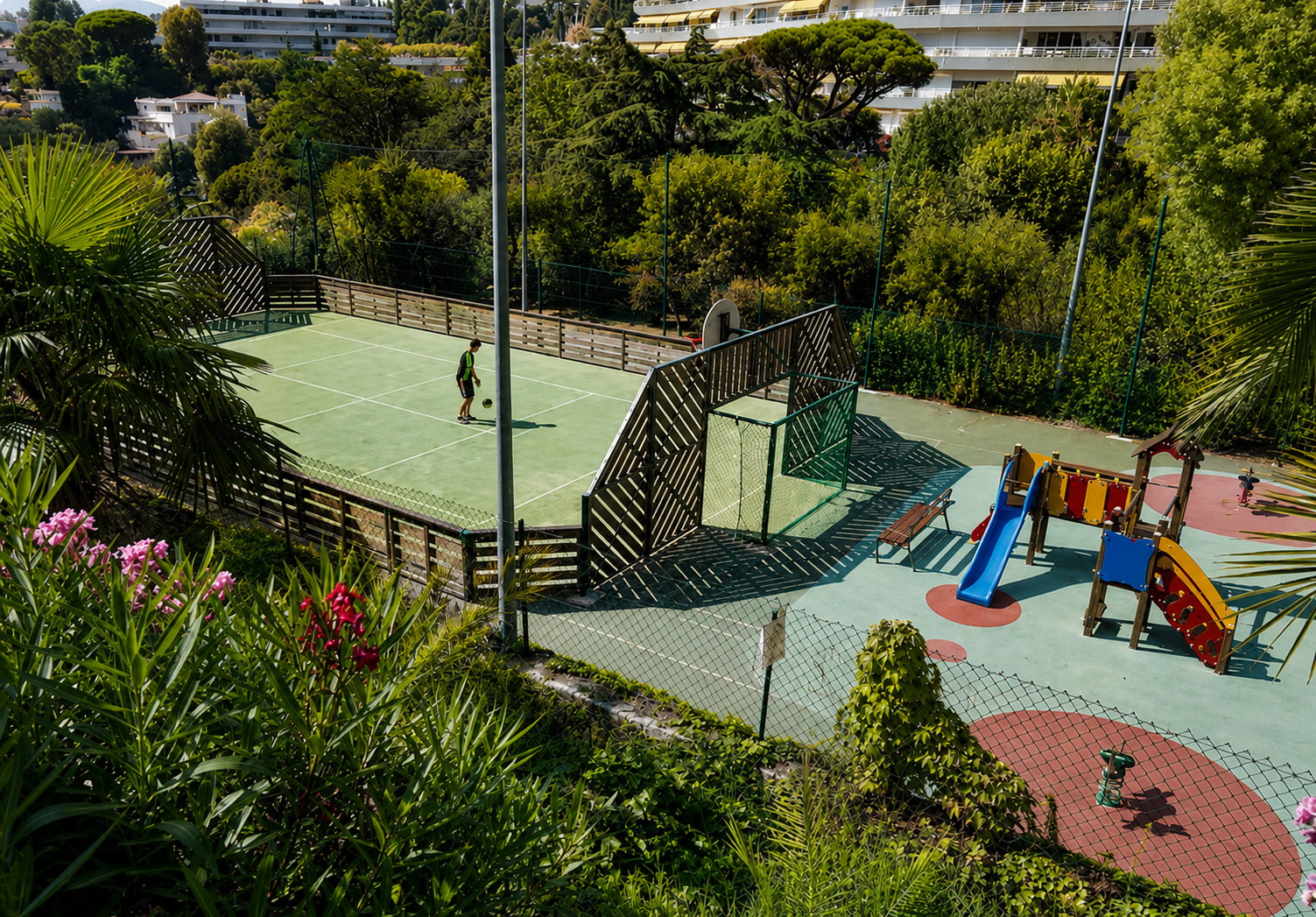 Terrains de jeu entourés de verdure avec une personne jouant au tennis et un espace de jeux pour enfants avec des toboggans.