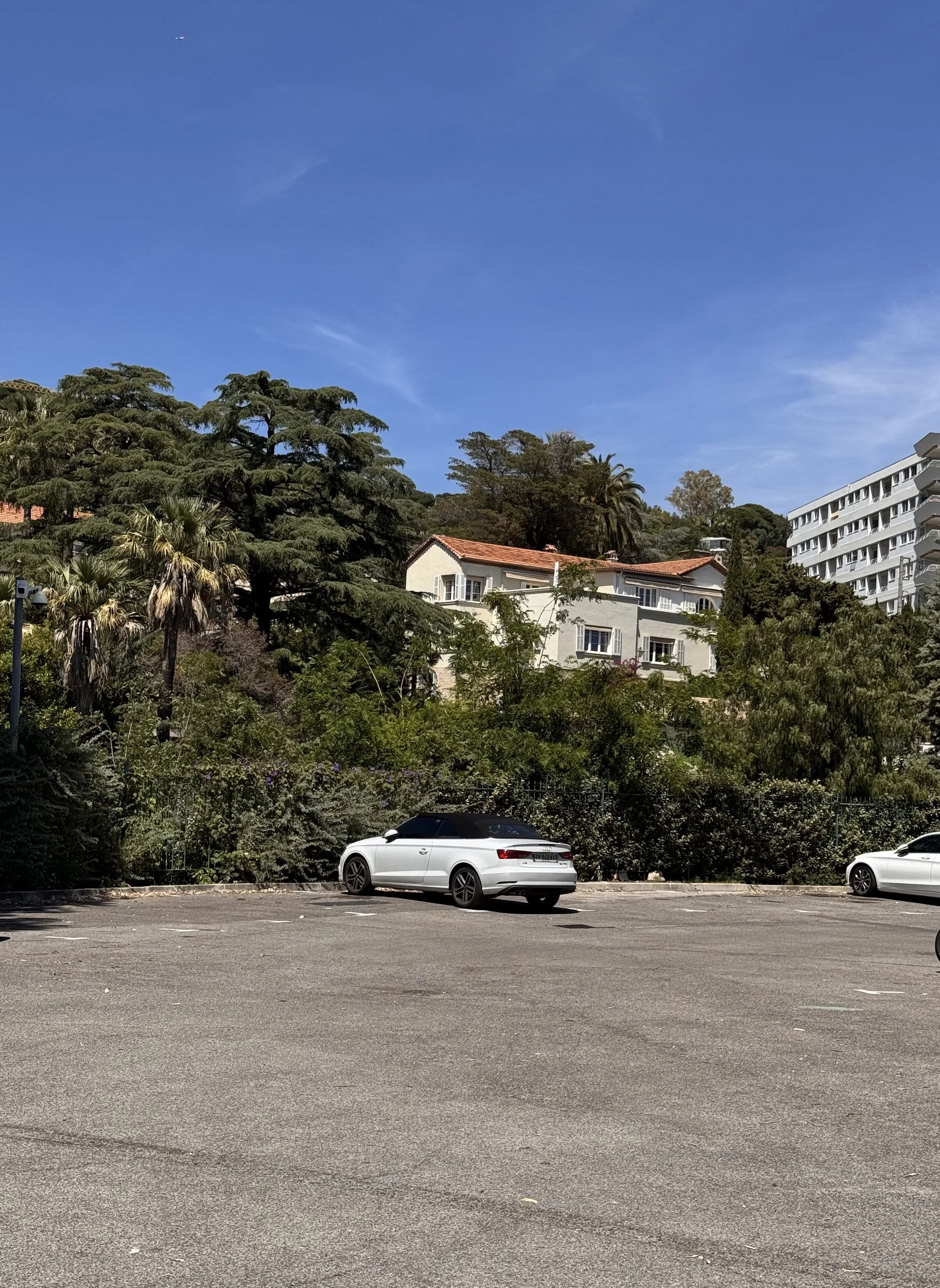 Un parking vide avec deux voitures blanches et un fond de bâtiments résidentiels et d'arbres, sous un ciel bleu clair.
