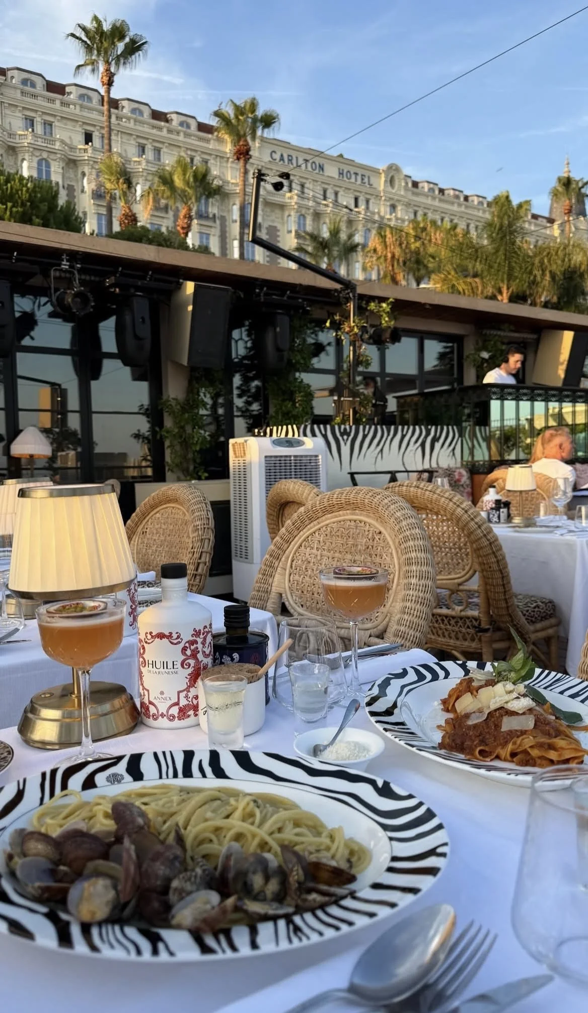 Repas italien avec pâtes et fruits de mer sur une table de restaurant en terrasse, ambiance en soirée, bâtiment El Carnon Hotel en arrière-plan avec palmier.
