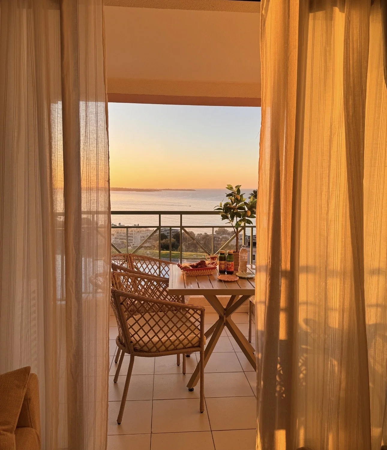 Balcon avec une table, des chaises en rotin, et une plante, vue sur la mer au coucher du soleil, vu à travers des rideaux transparents.