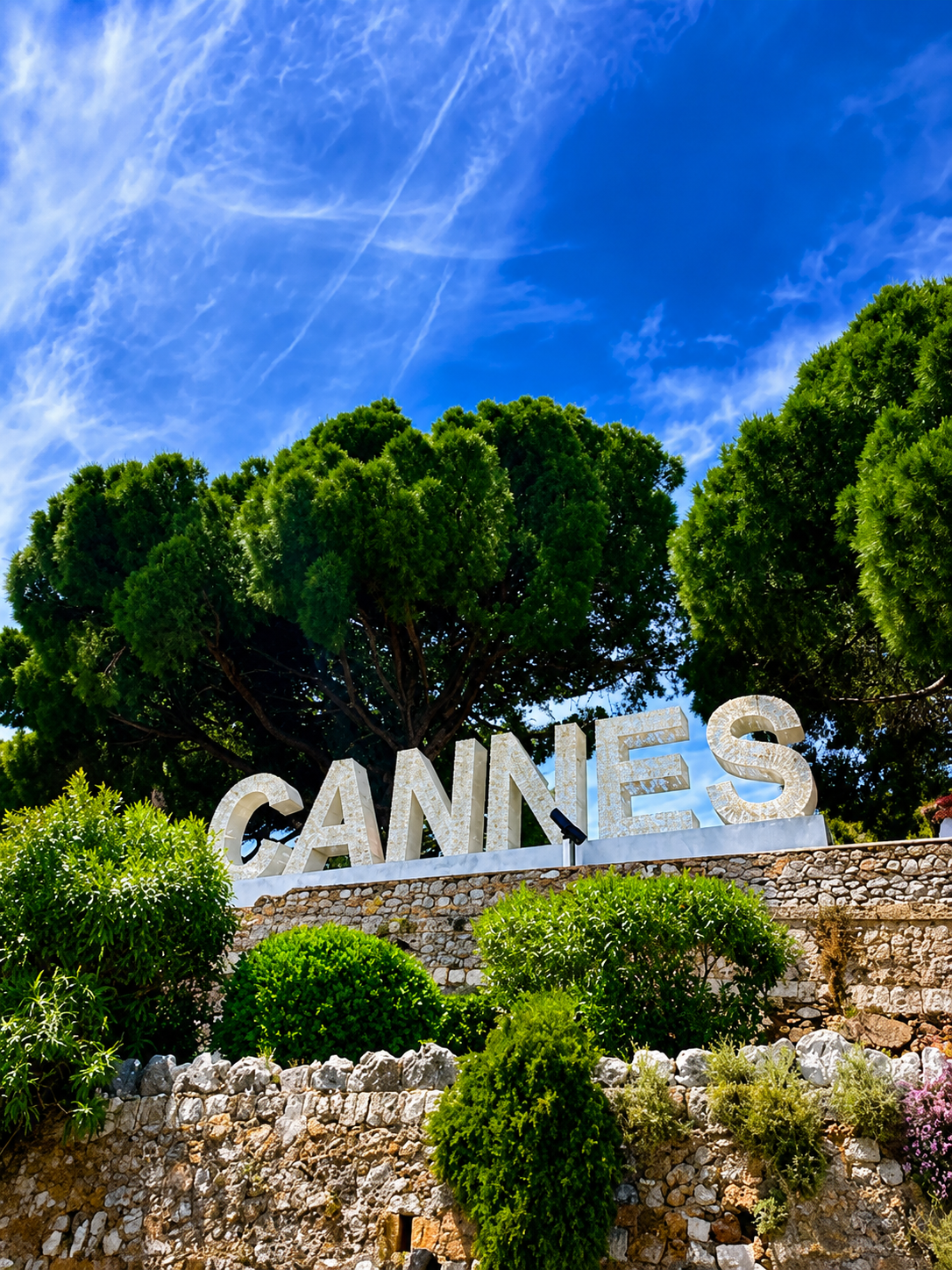 Le panneau lumineux 'CANNES' en lettres blanches géantes devant un arbre vert, sous un ciel bleu avec quelques nuages et une traînée d'avion.