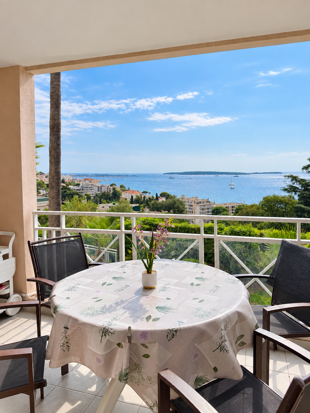 Balcon avec table ronde recouverte d'une nappe à motifs floraux, vase avec fleurs, vue sur la mer, bateaux et buildings, ciel bleu avec quelques nuages.