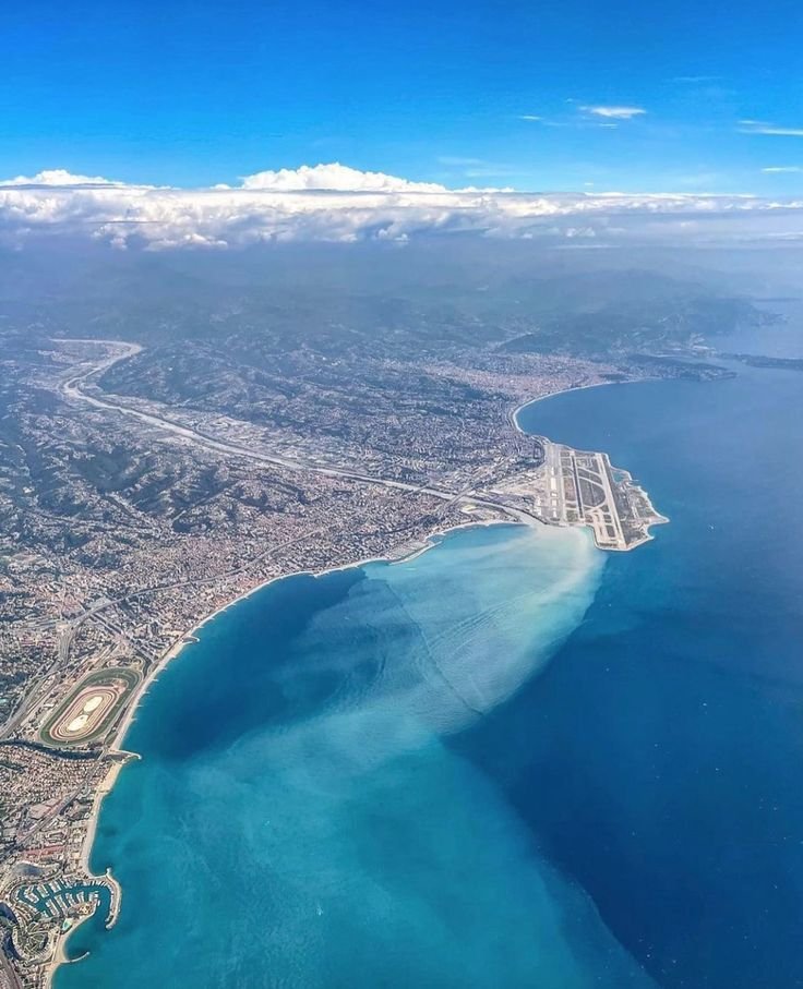 Vue aérienne d'une côte avec une plage, un port et une ville derrière, sous un ciel partiellement nuageux.