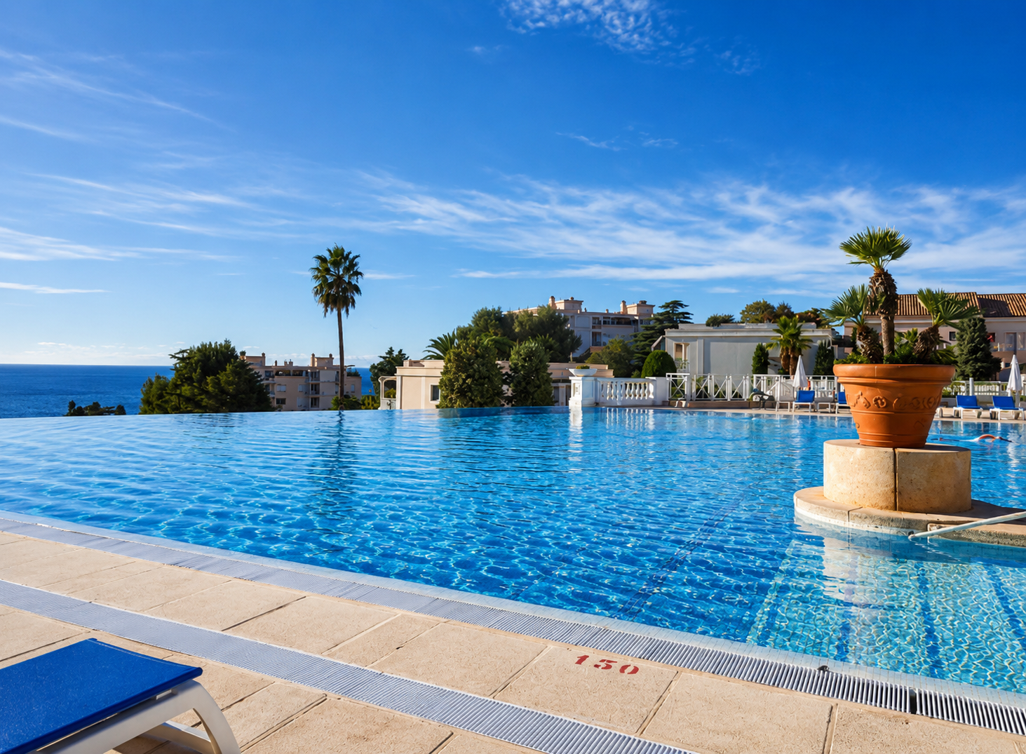 Piscine à débordement avec vue sur la mer, entourée de palmiers, terrasse et bâtiments résidentiels, ciel bleu et nuages légers.