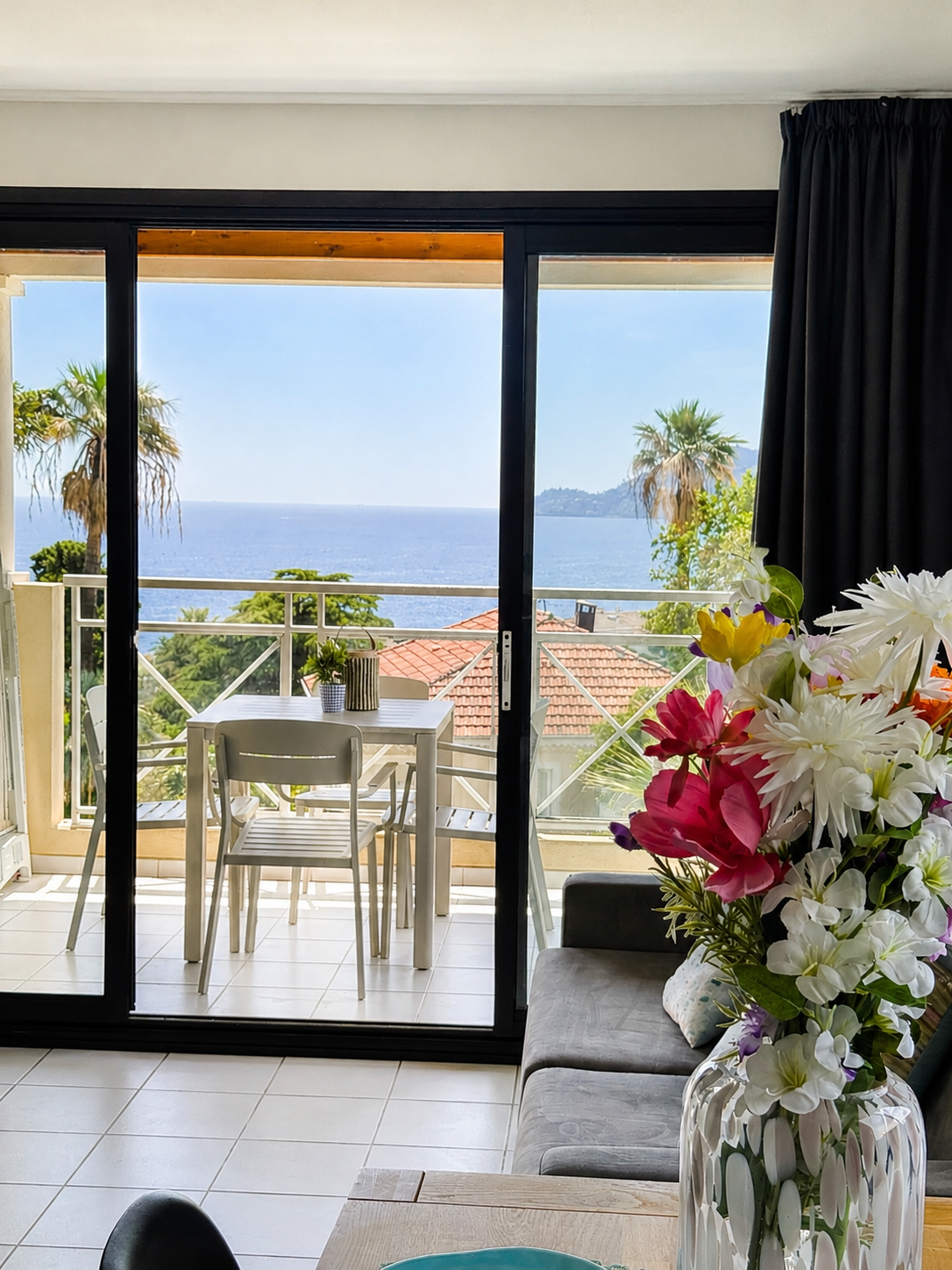 Vue intérieure d'un salon avec un canapé gris, une table basse et un grand vase de fleurs colorées, donnant sur un balcon avec une table et des chaises, vue sur la mer, palmiers et toits de maisons.