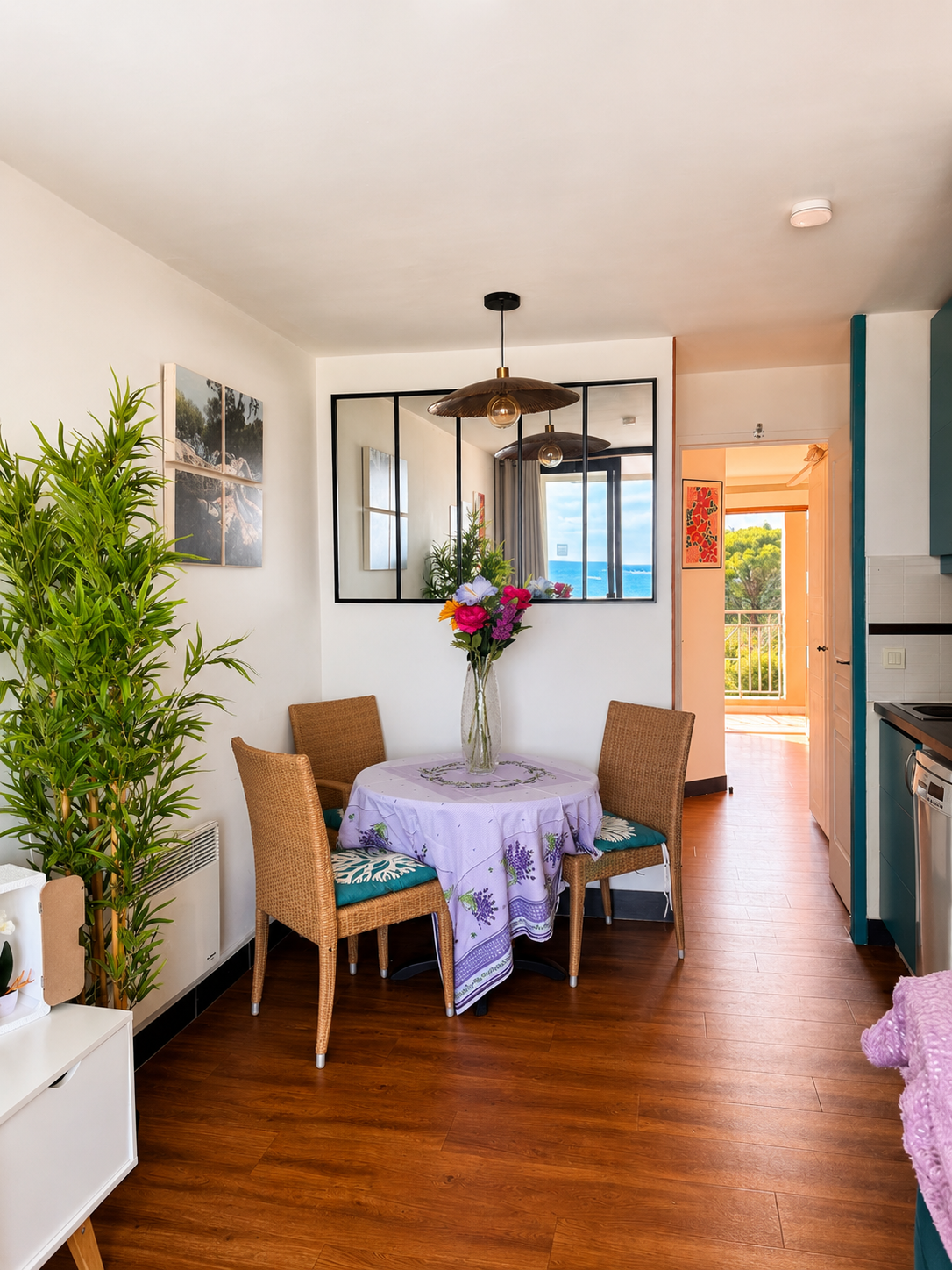 Intérieur d'une salle à manger avec un tapis rond, une table avec nappe lavande, un vase de fleurs, quatre chaises en rotin, plantes vertes et images sur les murs, vue vers un balcon avec ciel bleu.