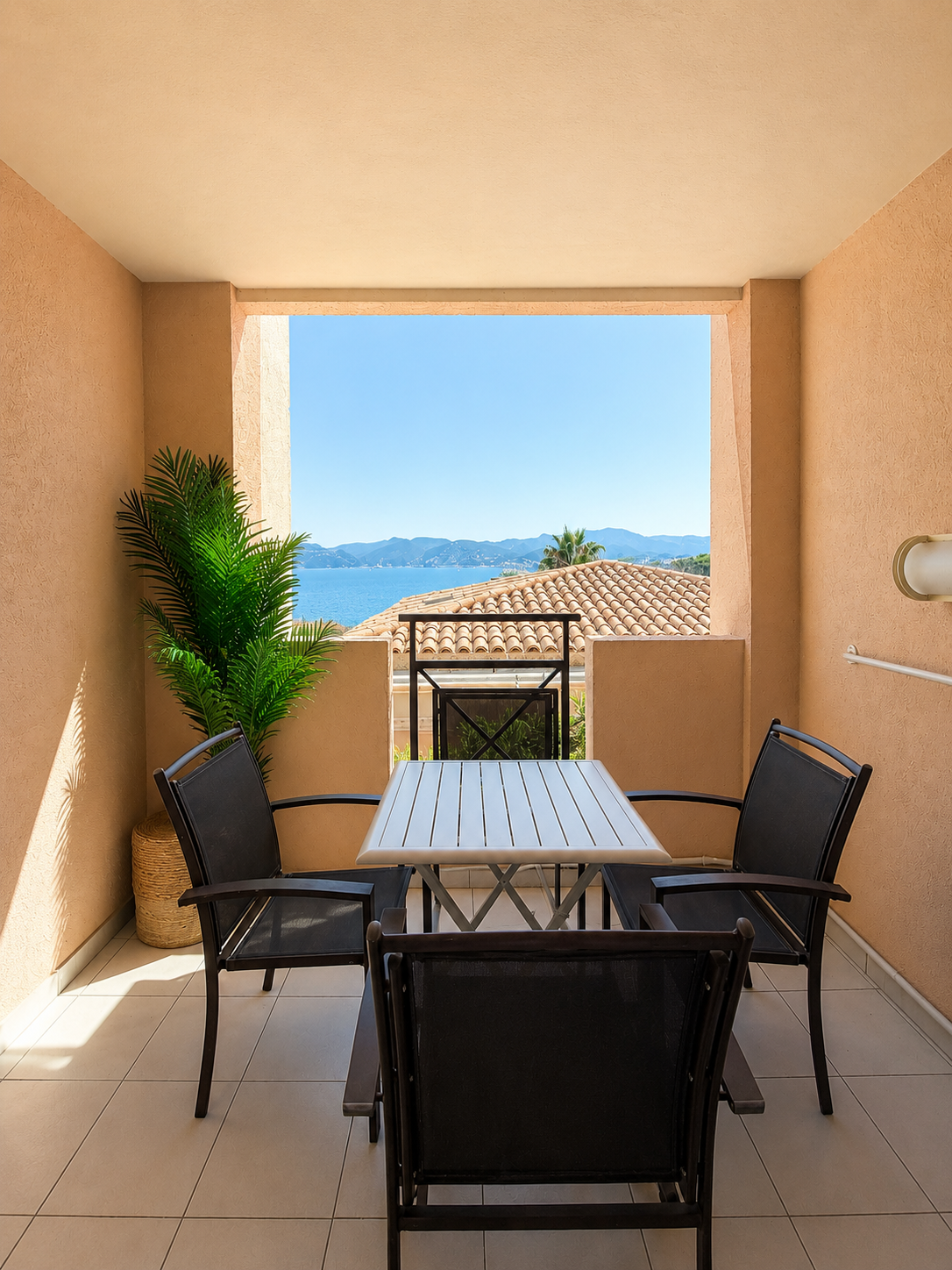 Balcon avec une table en bois et trois chaises noires, vue sur la mer et des montagnes au loin, avec un toit en tuiles en arrière-plan.