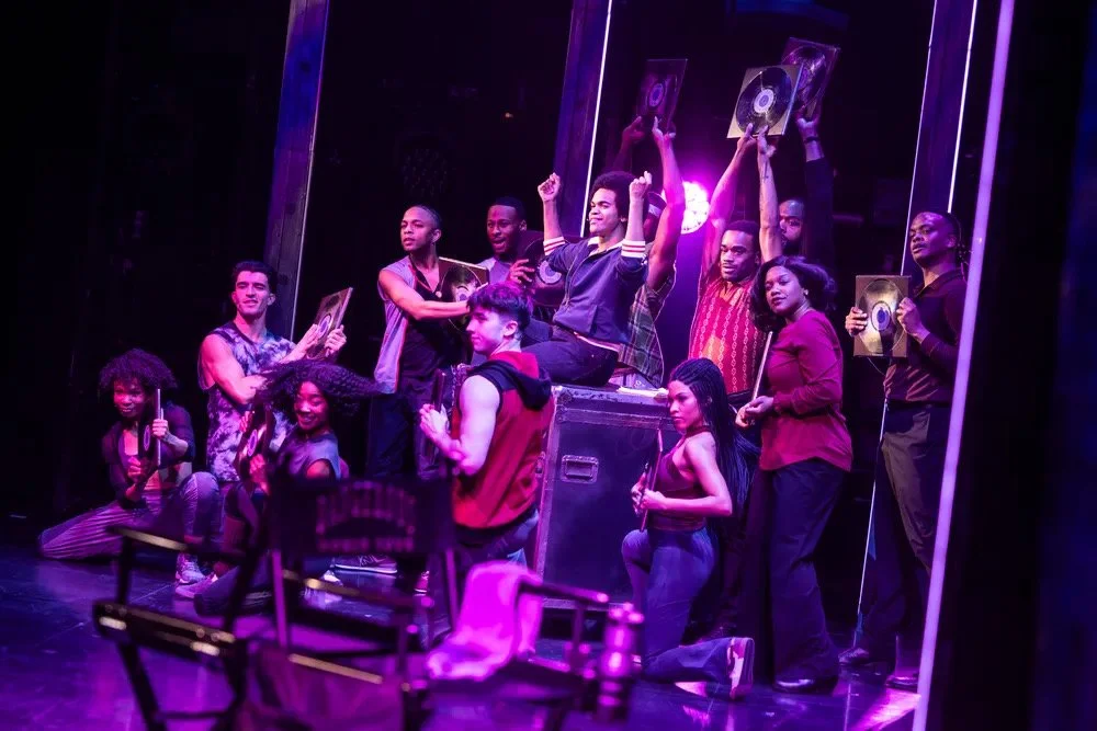Theatre cast performing on stage with some holding vinyl record covers, under purple stage lighting.