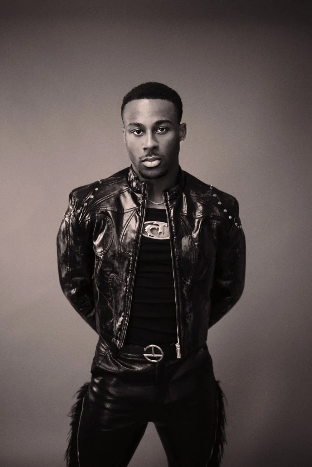 A young man with short hair wearing a black leather jacket with metal studs, a black T-shirt with a metallic Superman logo, black pants with a silver belt, posed against a plain, neutral background.