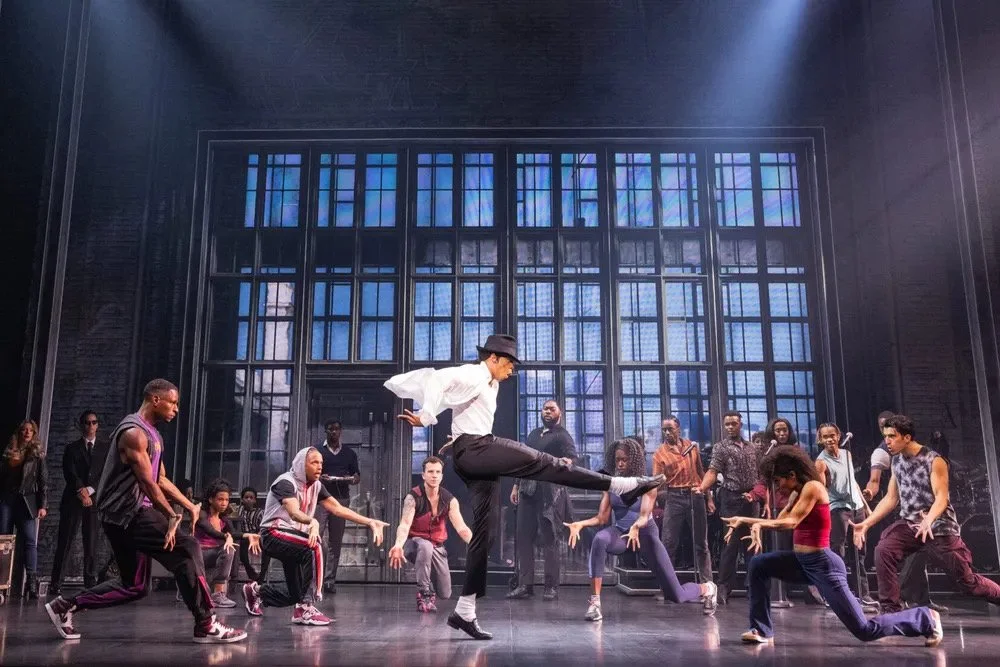 Stage scene with a male dancer in a white shirt and black hat performing a high kick, surrounded by diverse dancers in casual attire, set against a backdrop of large blue-tinted windows.